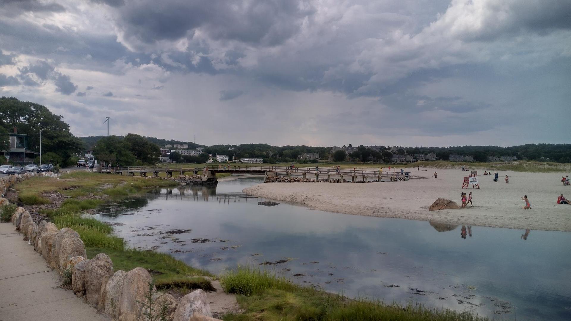 Overcast day over a river, sandy beach, and a bridge with people.