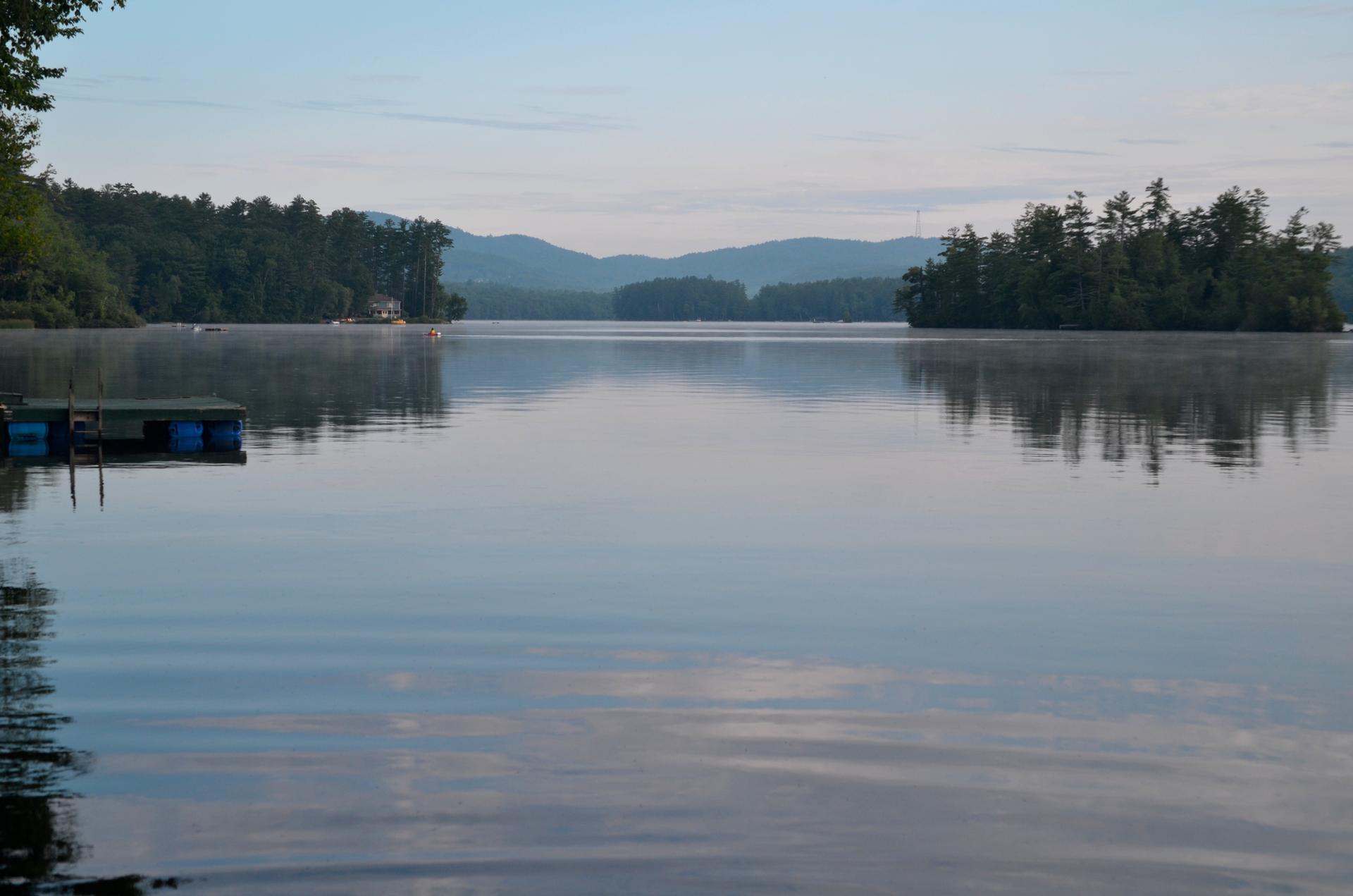Calm lake with forested shores, distant mountains, and a small dock under soft light.