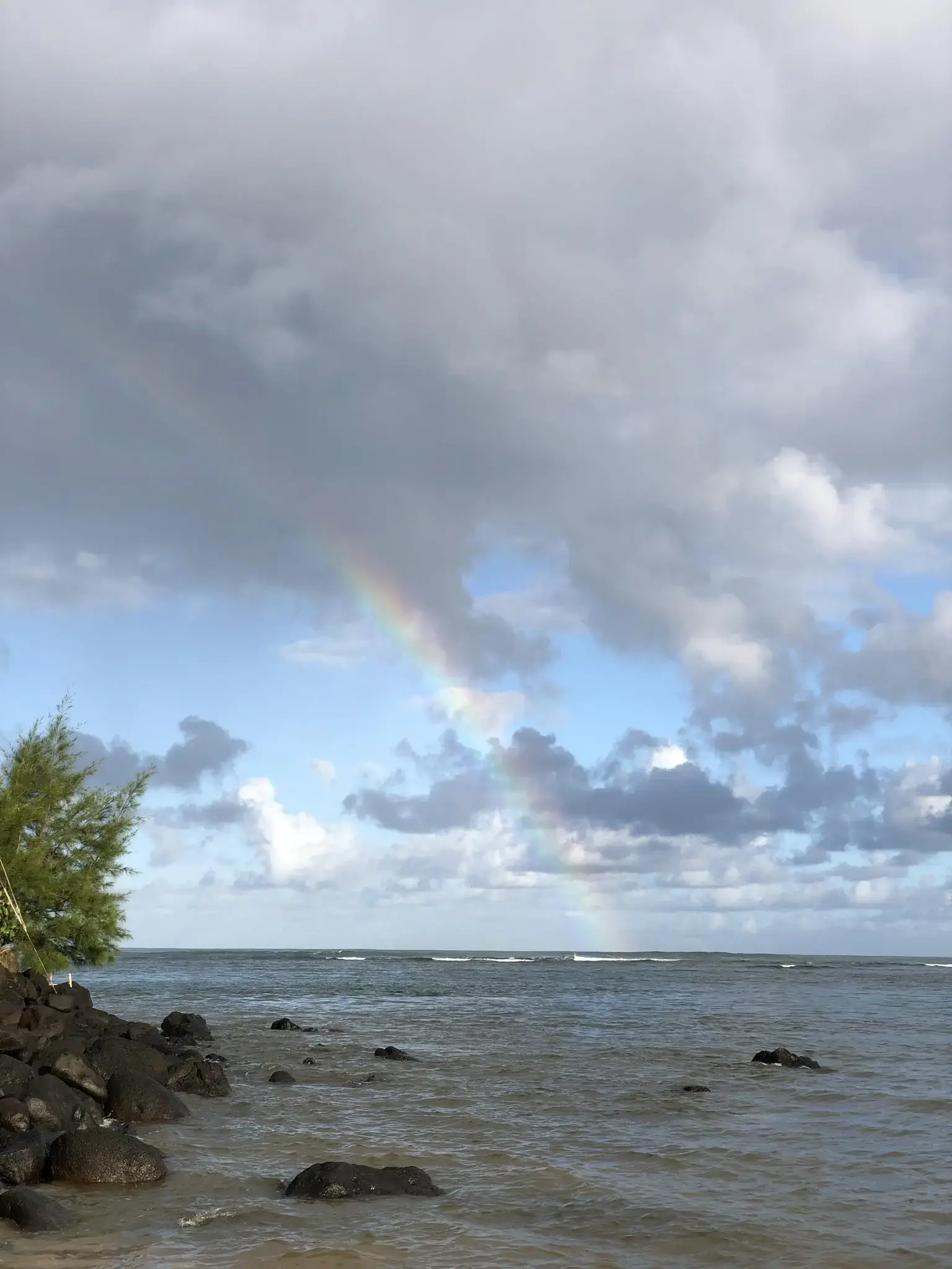 Overcast sky with a rainbow over a rocky ocean shore and breaking waves.