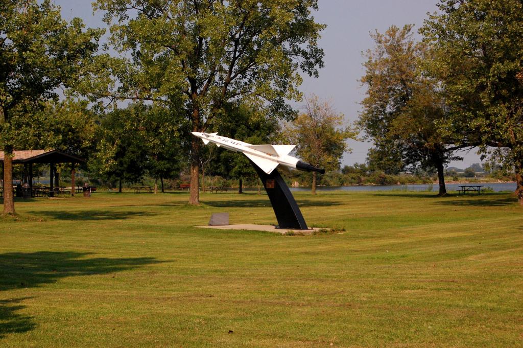 A white missile on a black stand in a sunny, grassy park with trees.