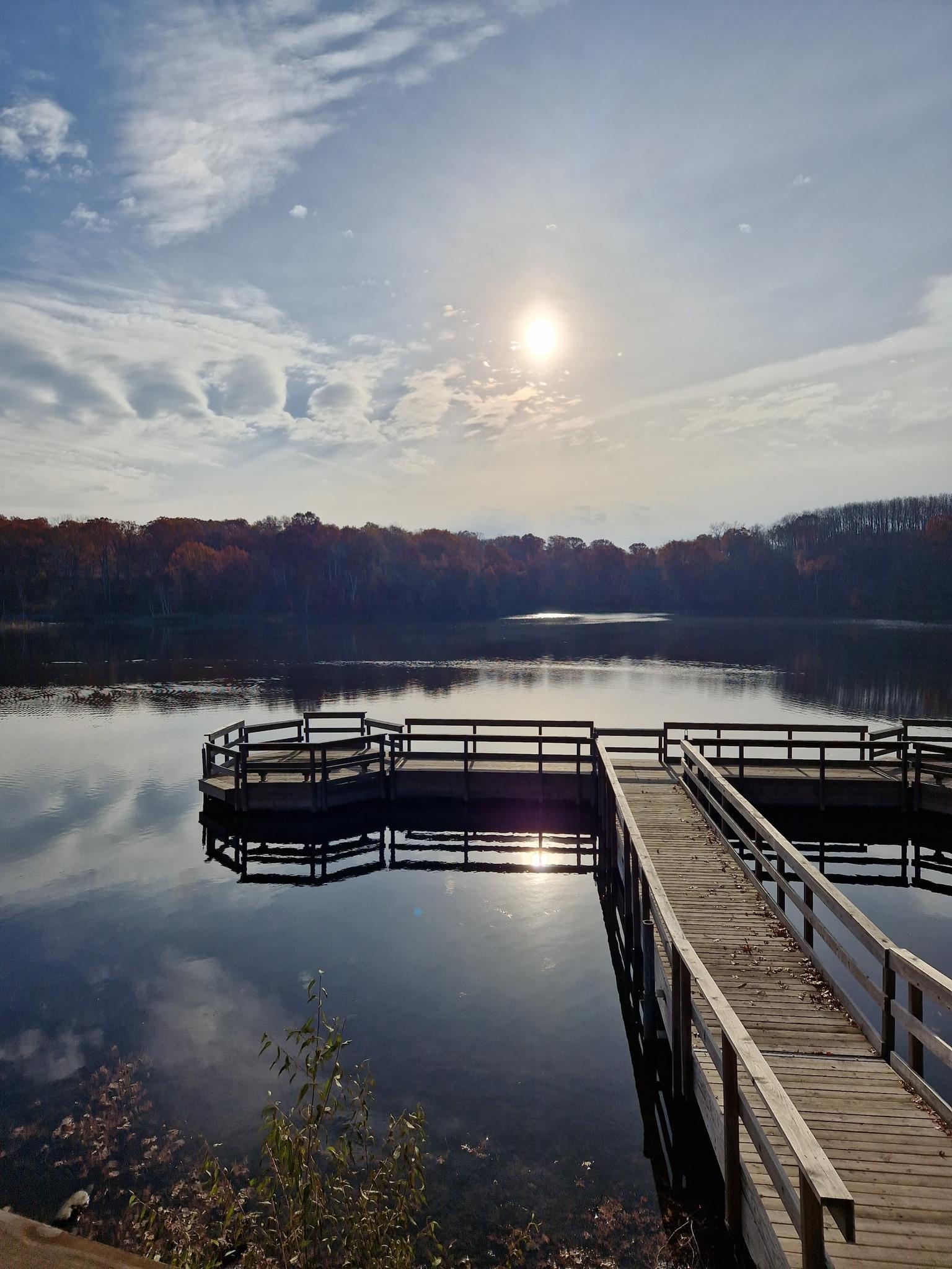 Wooden pier extending into a calm lake, sun shining brightly in a cloudy sky.