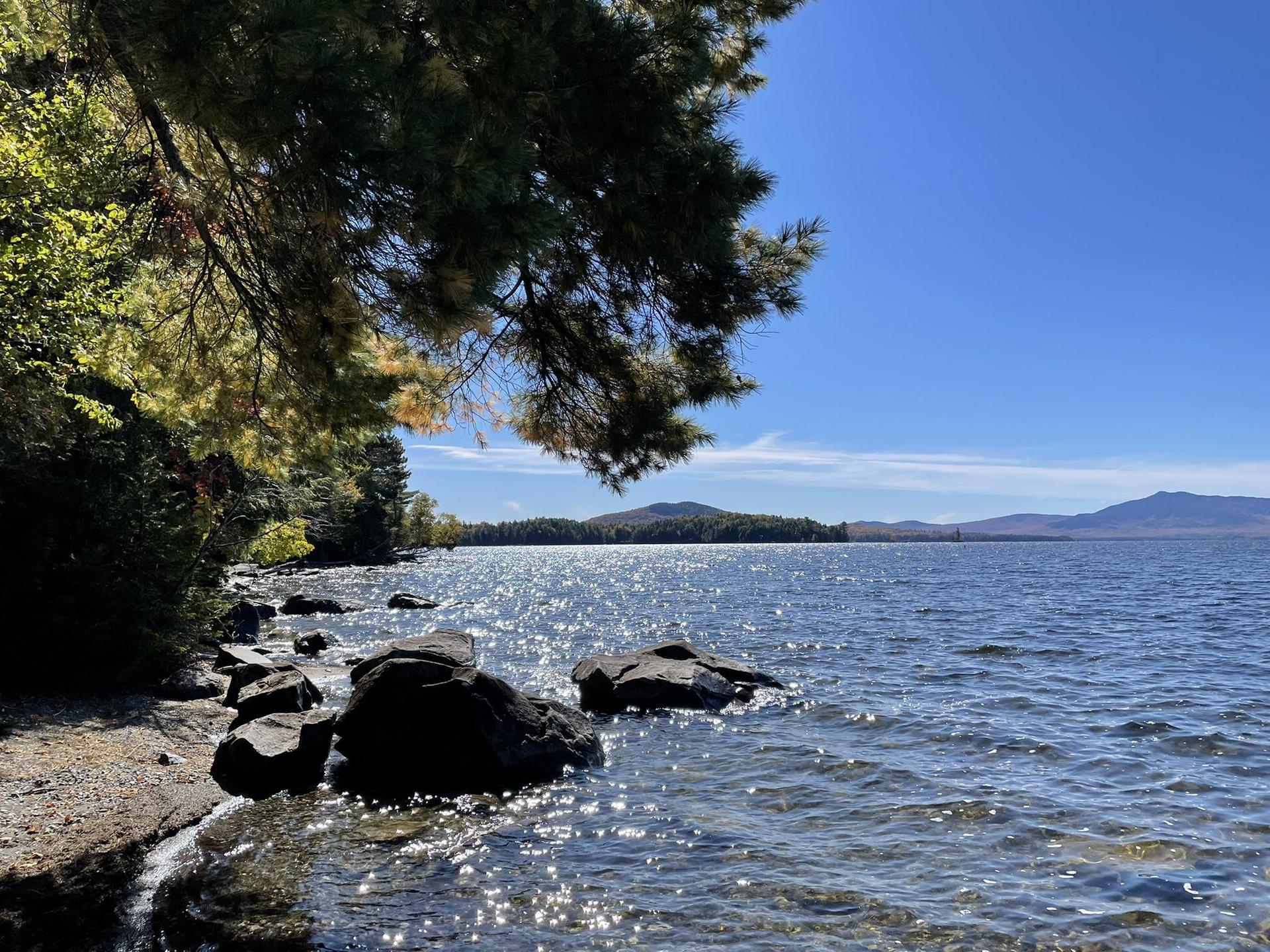 Sunny lake with sparkling water, rocky shore, and distant mountains under blue sky.