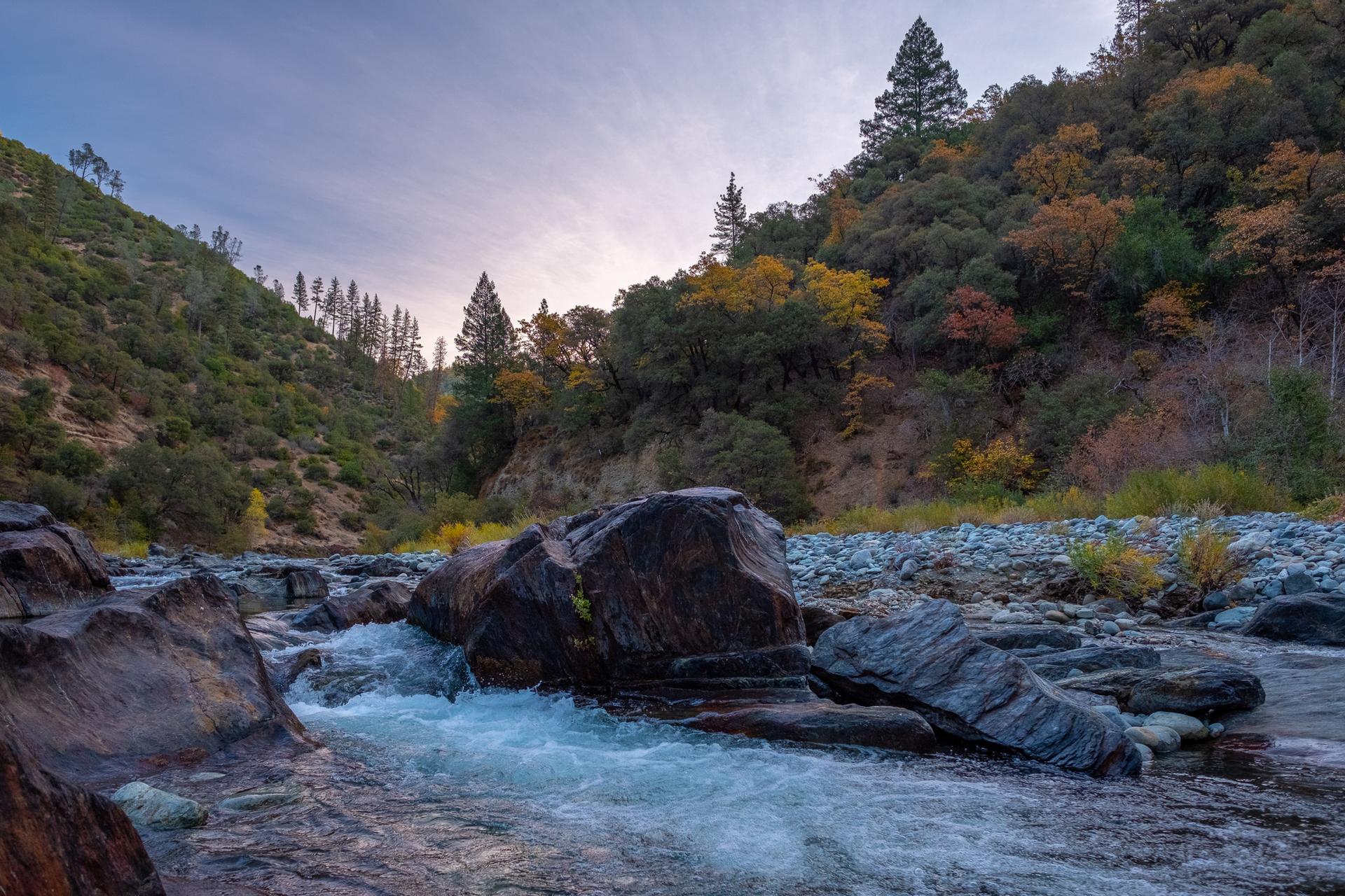 Rushing river over dark rocks, surrounded by forested hillsides under a cloudy sky.