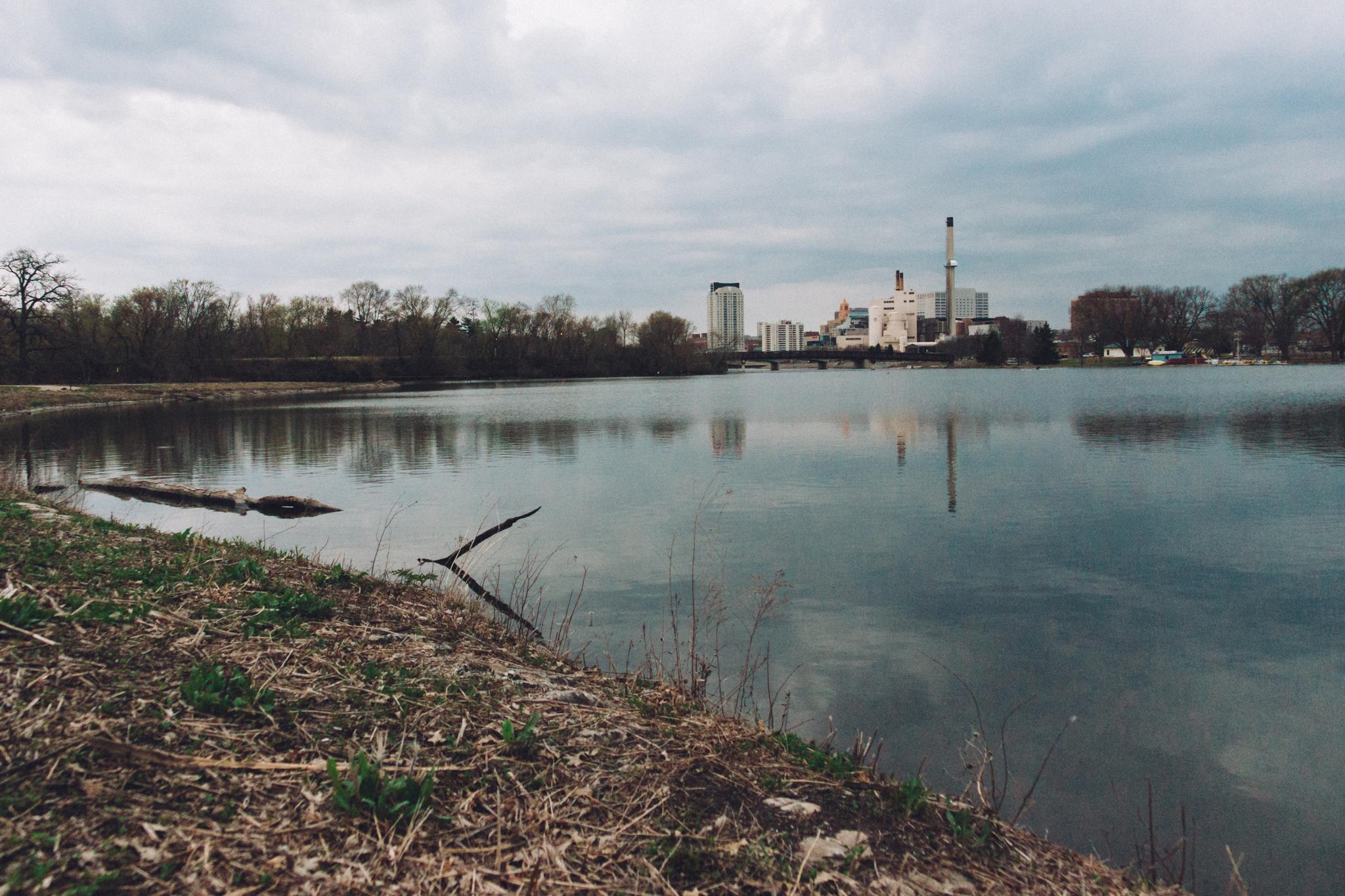Industrial buildings reflected in calm water under a cloudy sky.