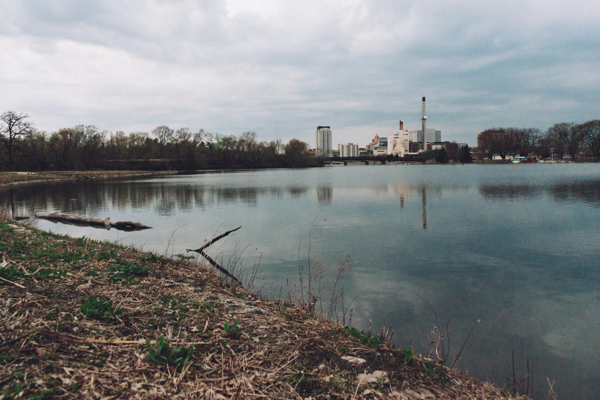 Industrial buildings reflected in calm water under a cloudy sky.