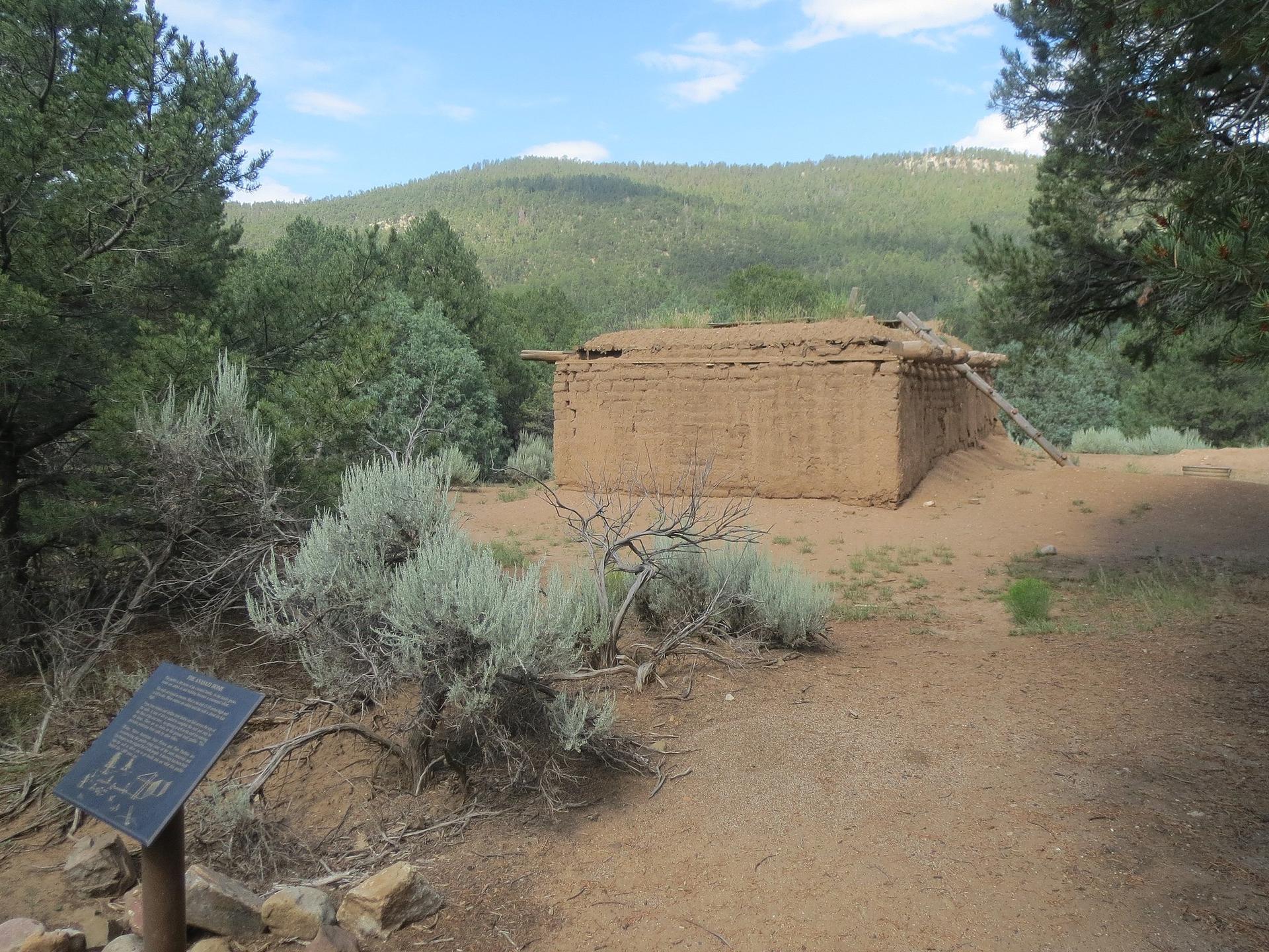 An old adobe structure in a dry, forested landscape under a bright sky.