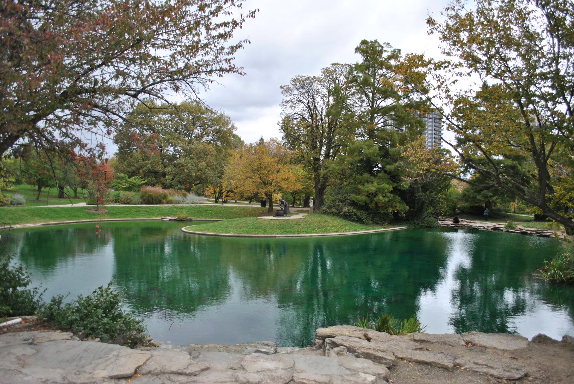 Green pond with a small island, surrounded by trees under an overcast sky.