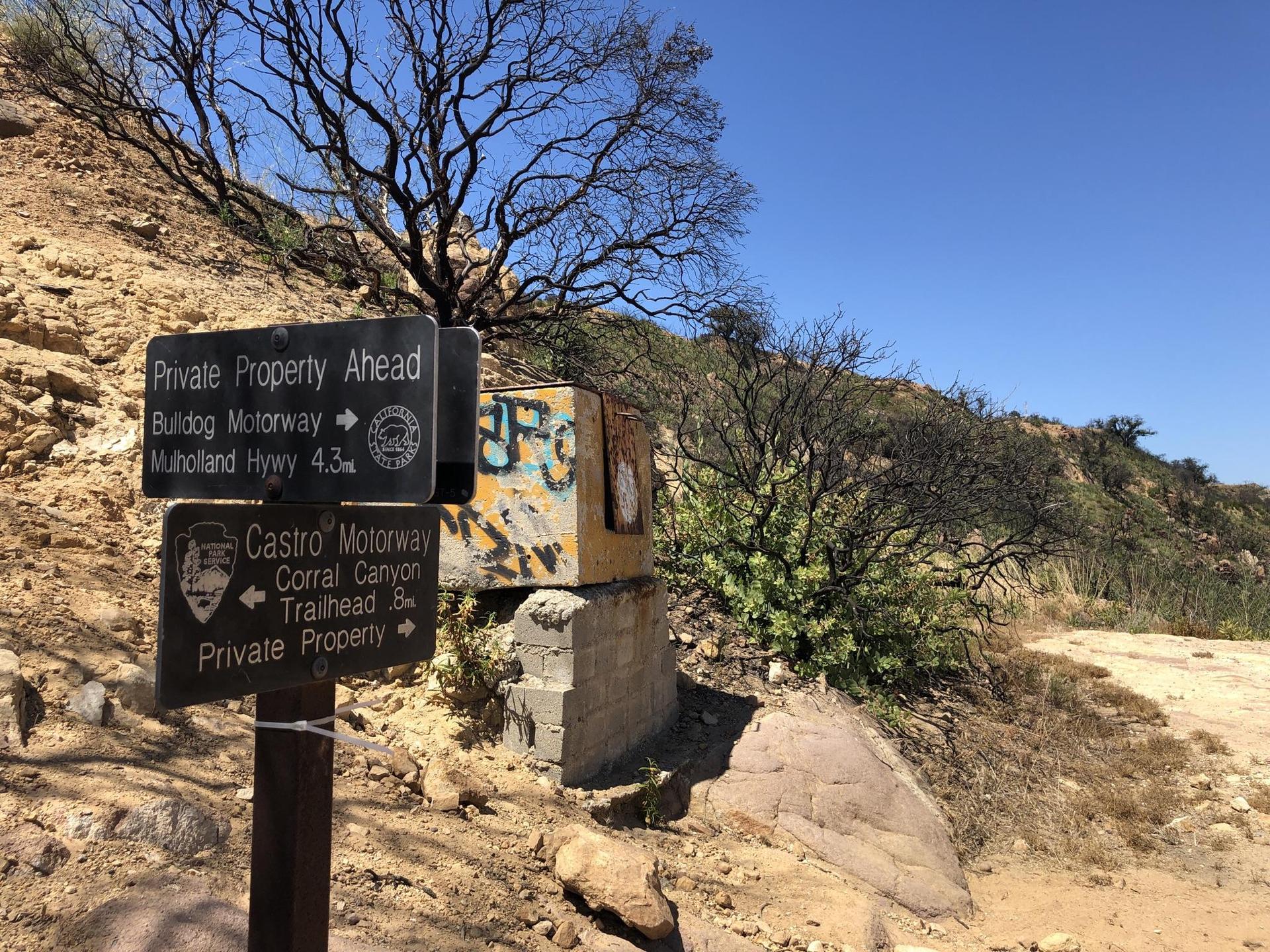 Trail signs on a post next to a graffiti-covered box on a hillside.