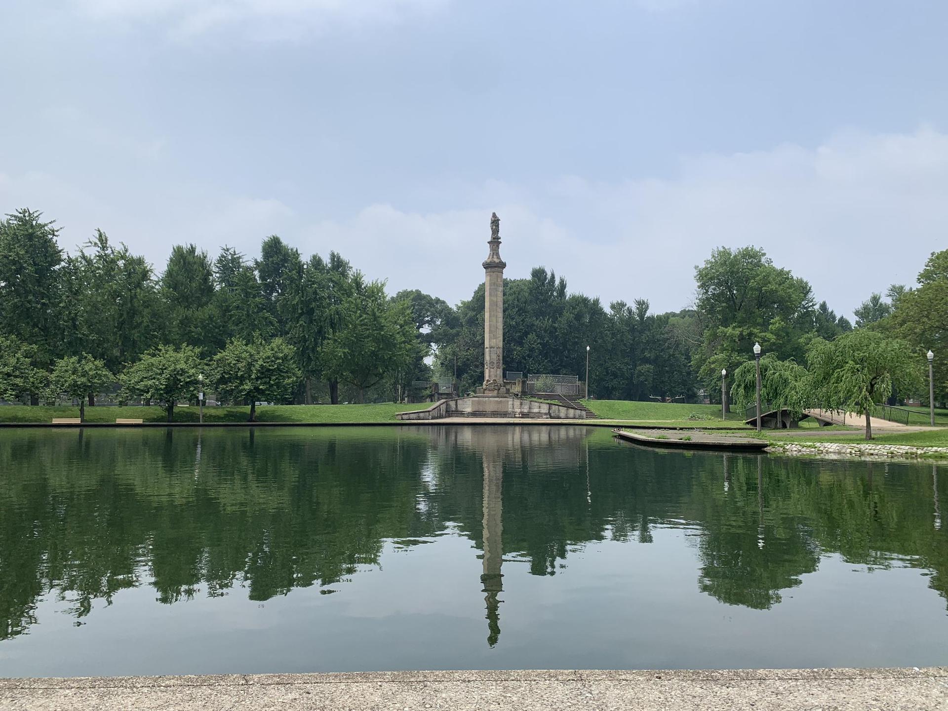 Tall stone monument reflected in a calm lake, surrounded by green trees.