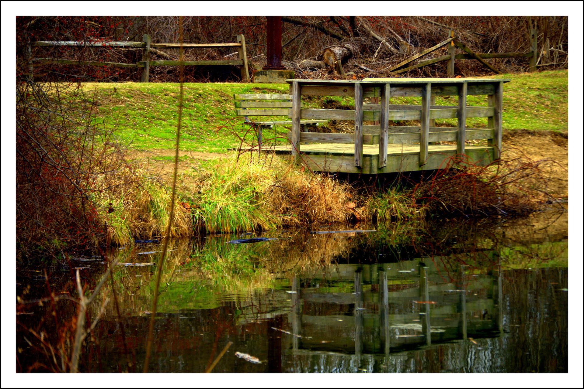 A small wooden bridge spans a dark pond, reflecting its structure and green banks.