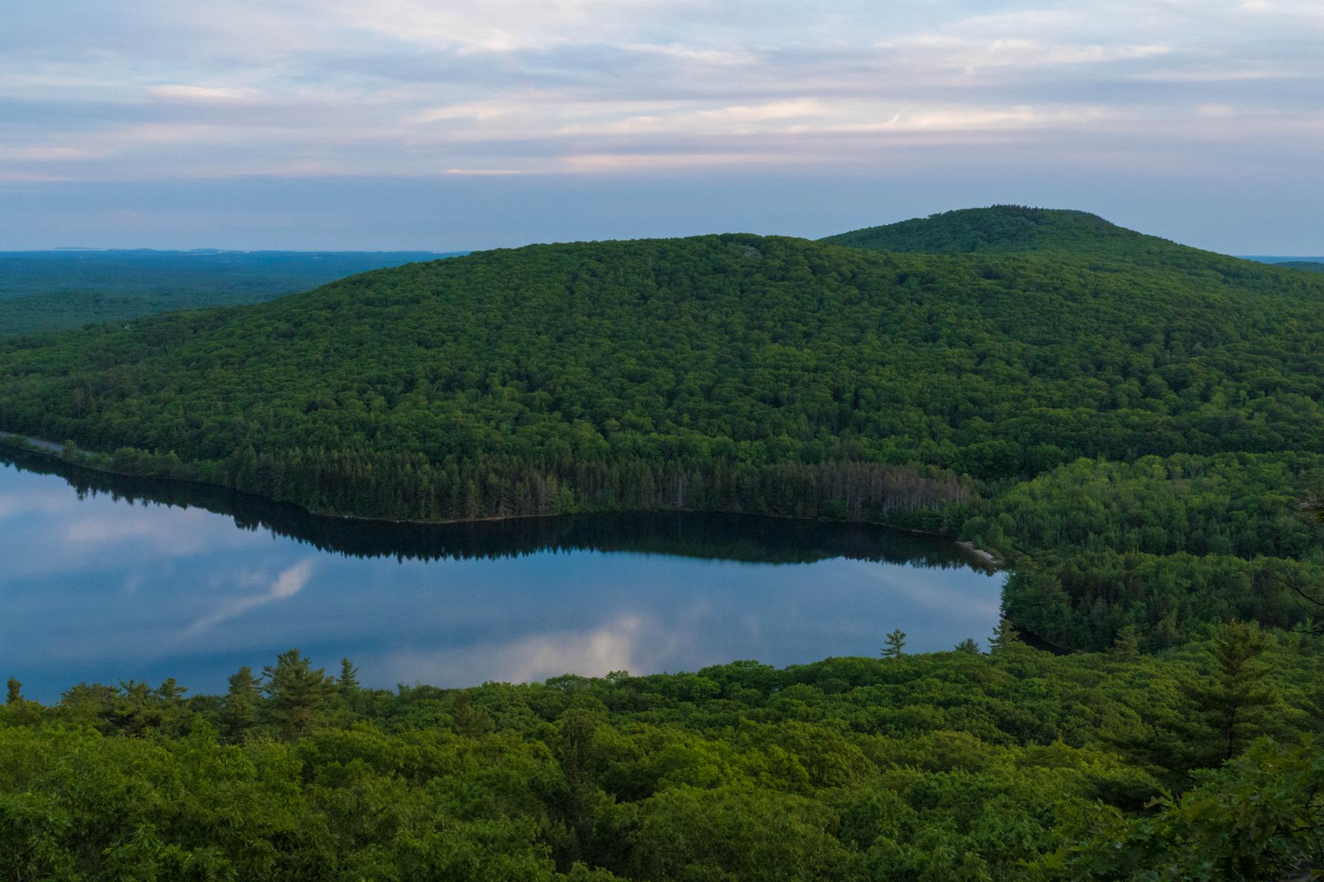 Overhead view of a calm lake reflecting cloudy sky, surrounded by green forests.