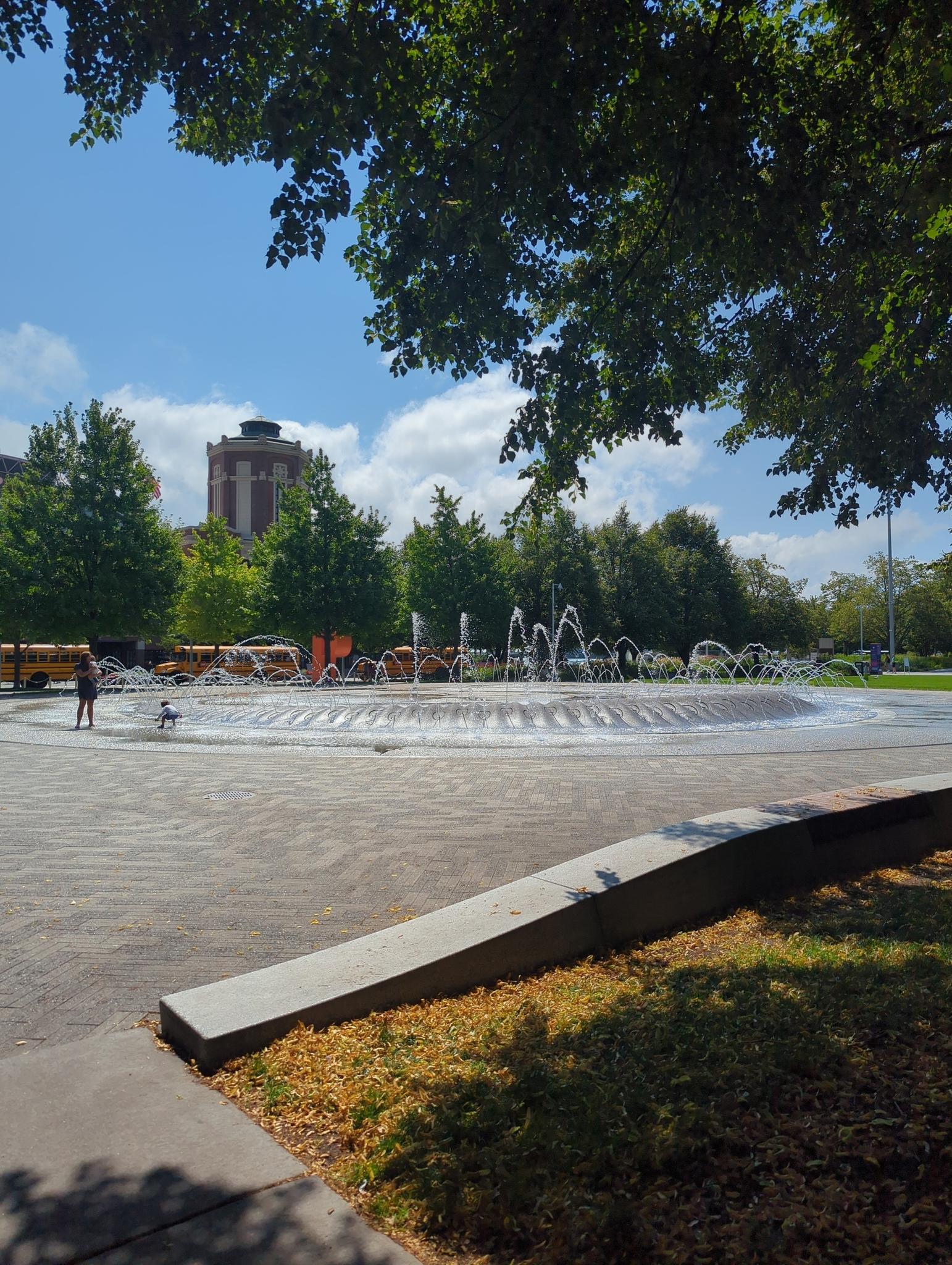 People near a large circular fountain in a park on a sunny day.
