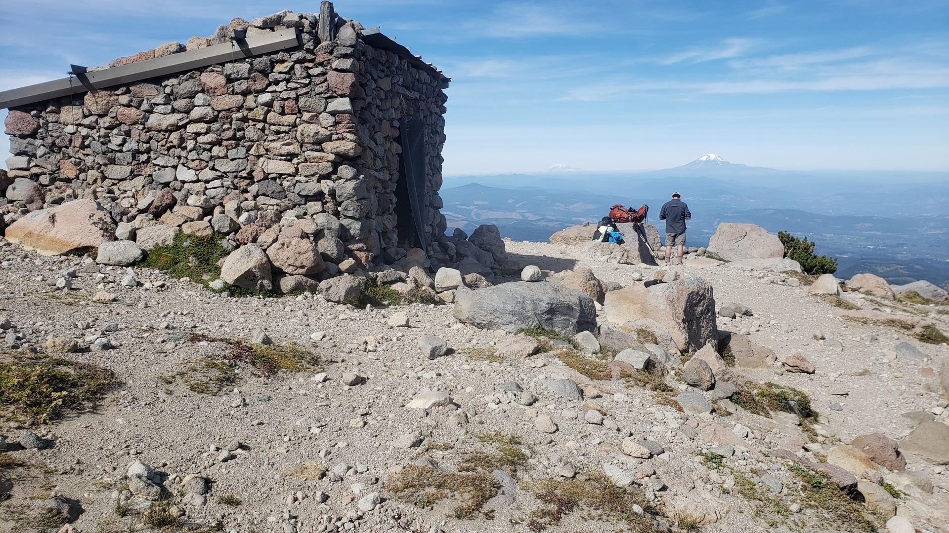 Two hikers stand near a stone shelter on a rocky mountain summit, clear day.