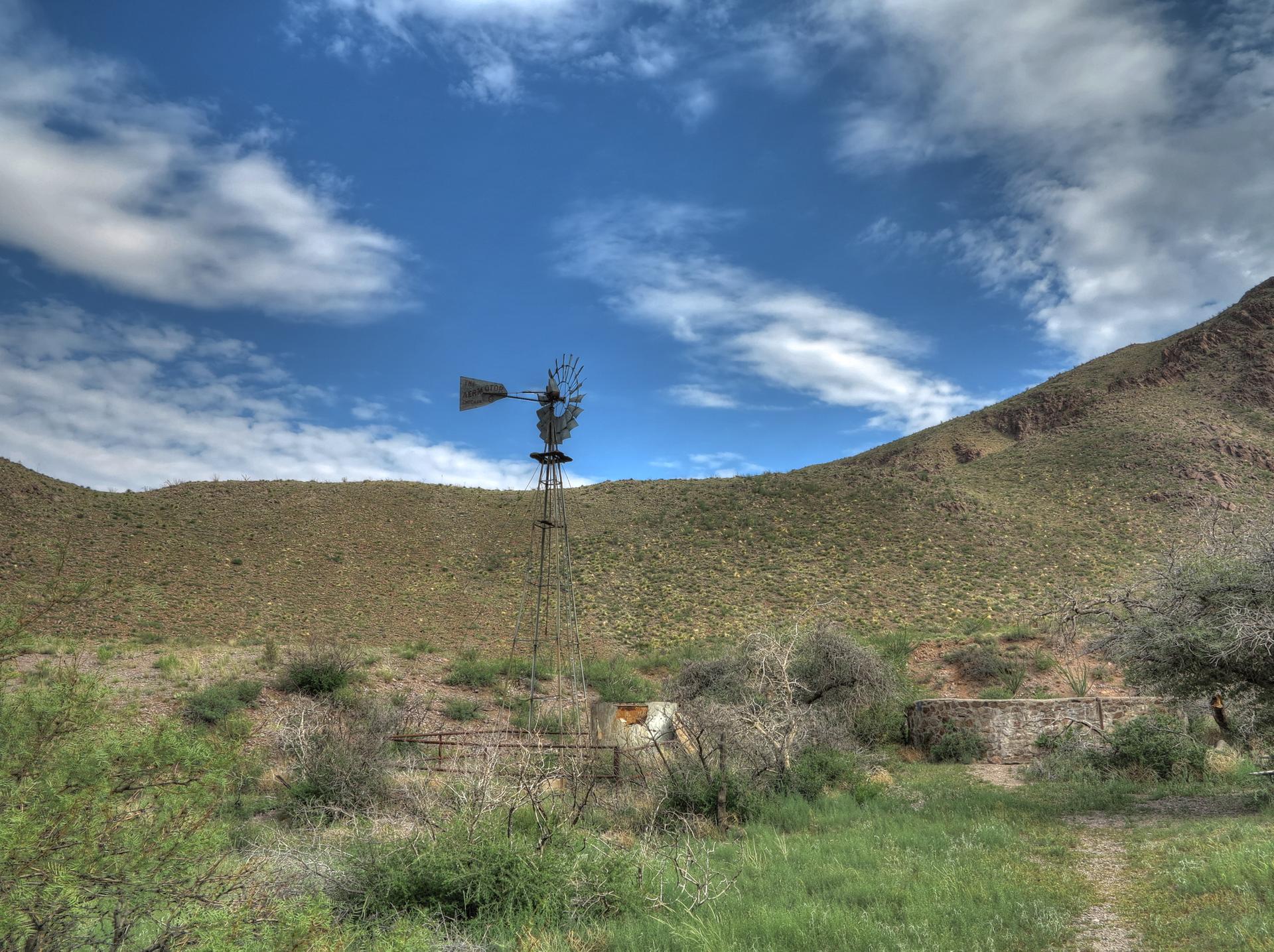 An old windmill stands in a green, hilly landscape under a bright, cloudy sky.
