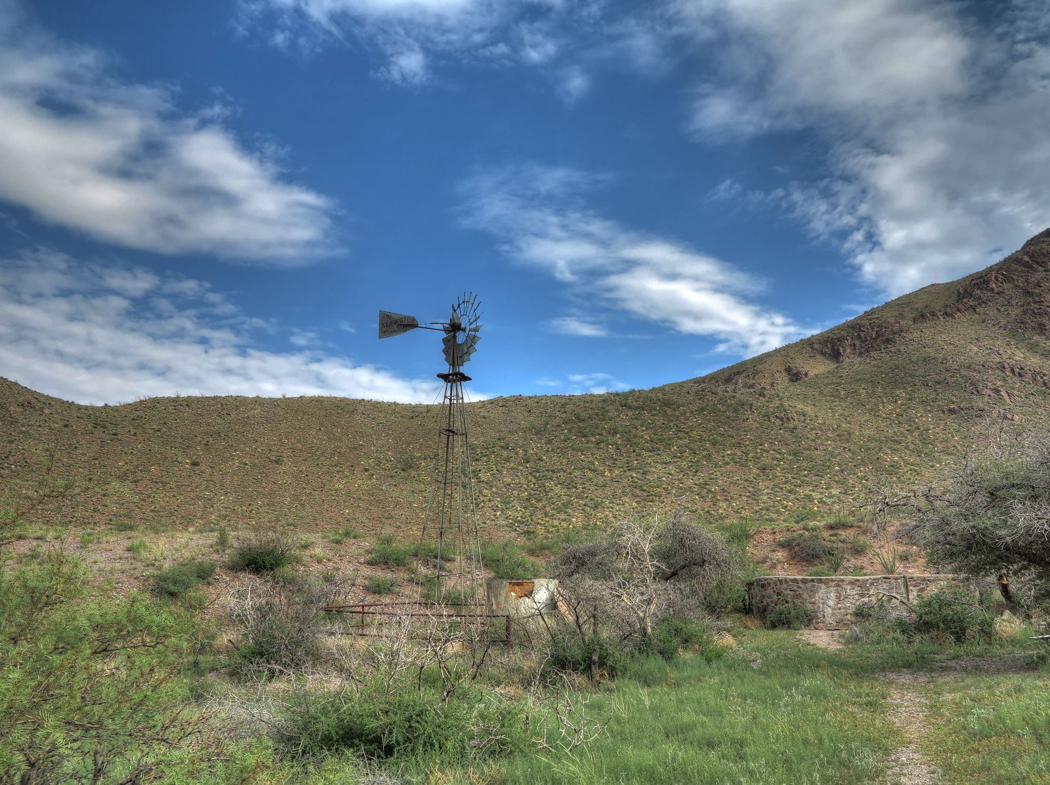 An old windmill stands in a green, hilly landscape under a bright, cloudy sky. - Bar Canyon