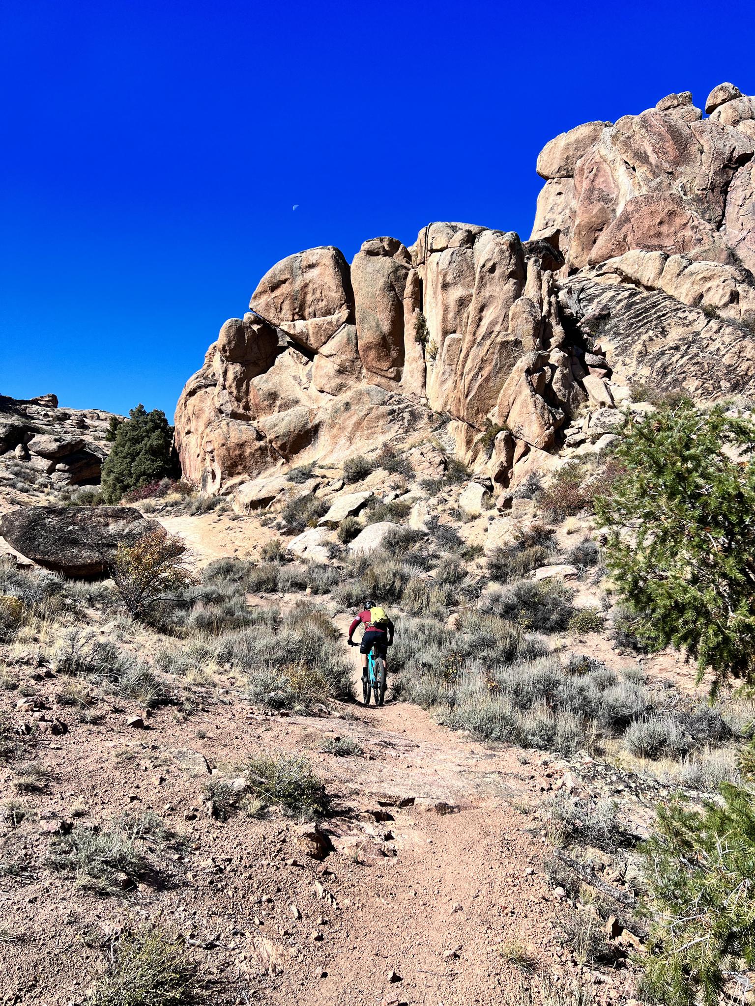 Mountain biker on a dirt path in a sunny, rocky, arid landscape.