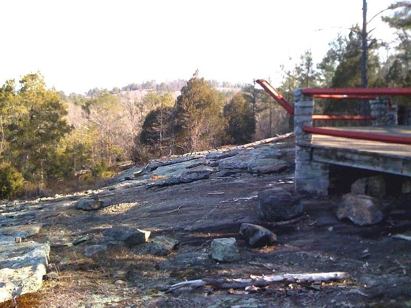 Rocky outcrop with a wooden viewing platform and distant trees under bright sky.