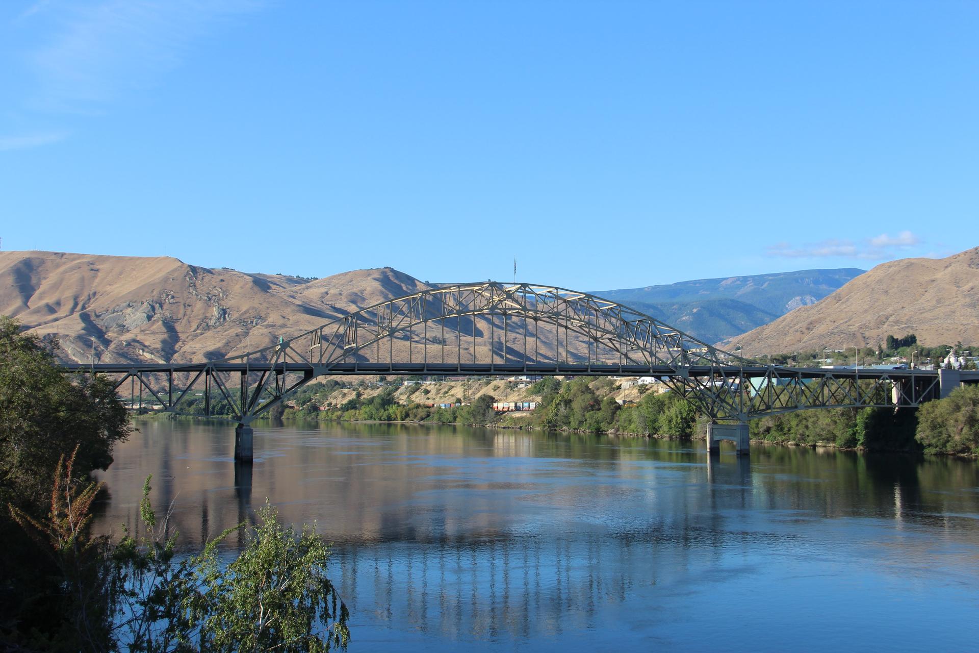 Large metal bridge spanning a wide river with mountains and trees under clear skies.