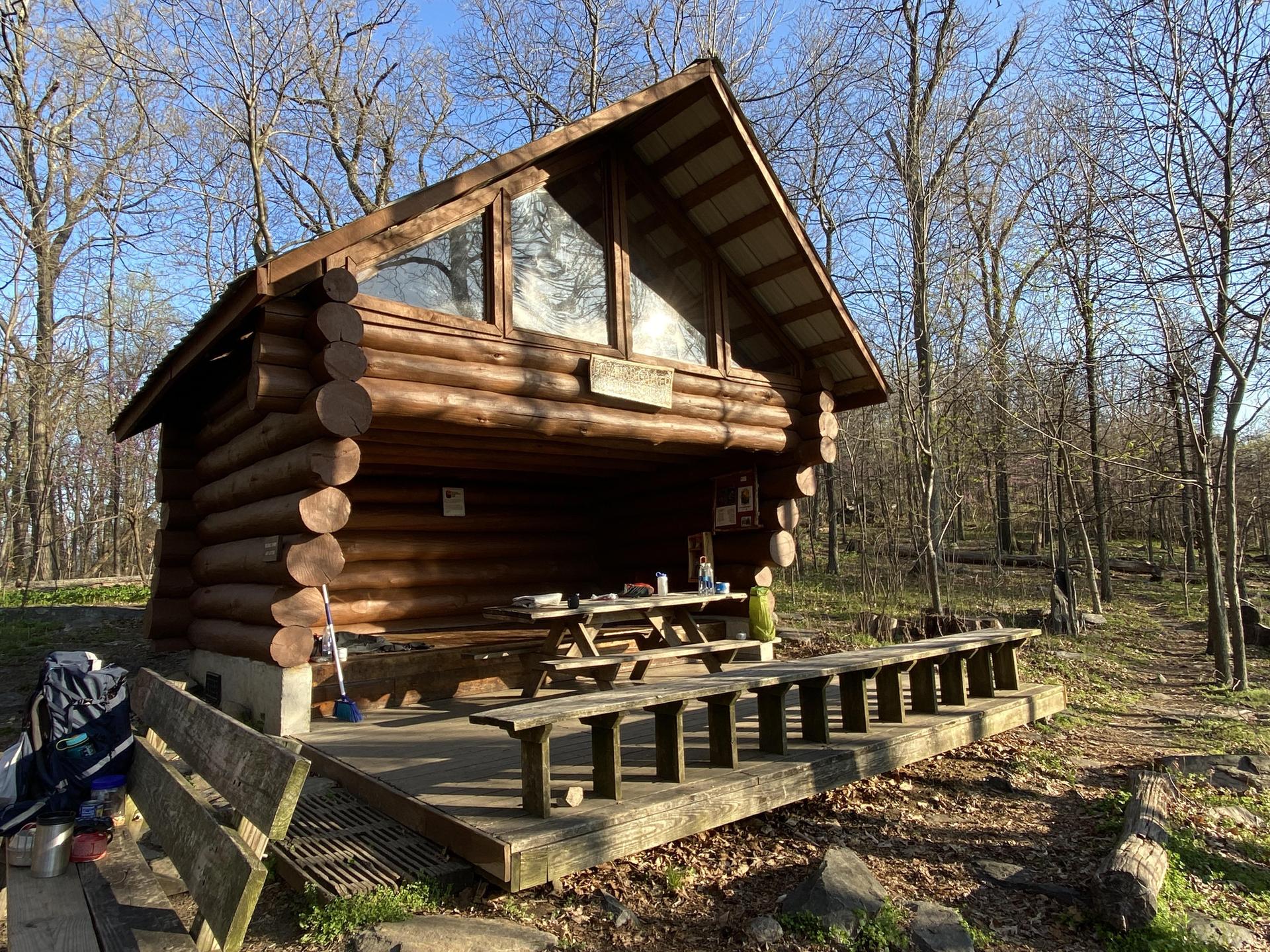 A rustic log cabin shelter in a sunny forest clearing with a backpack.