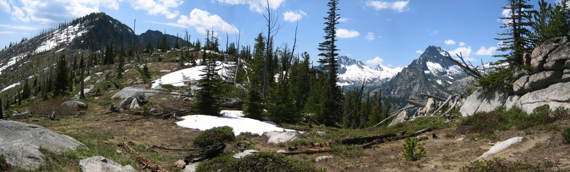 Sunny panoramic mountain landscape with snow patches, pine trees, and distant peaks.