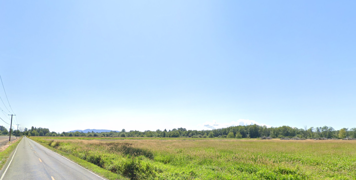 A long road next to a vast green field under a clear blue sky.