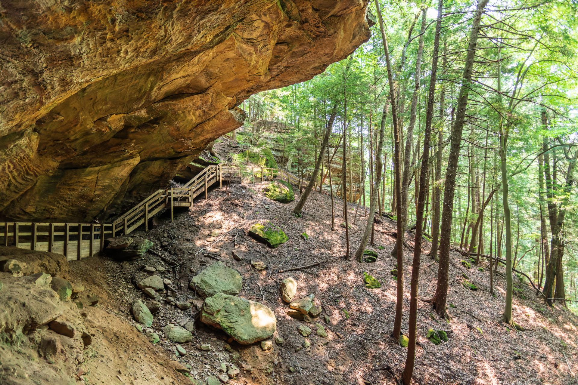 Wooden stairs and boardwalk under a large rock overhang in a sunlit forest.