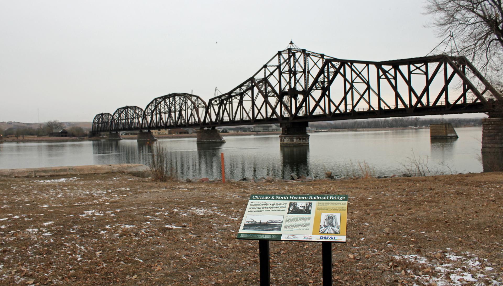 Large metal truss bridge over a river, with an informational sign in foreground.