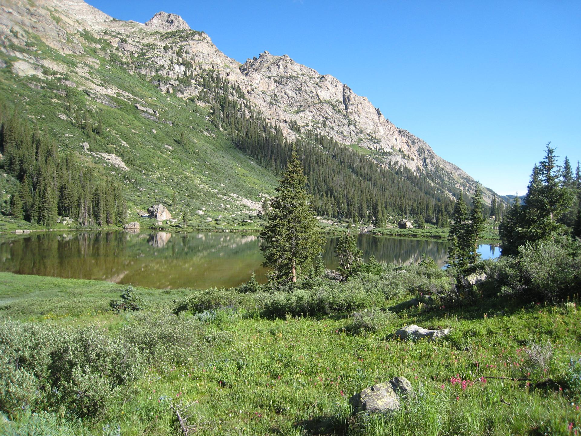 A serene mountain lake reflects rocky peaks and pine forests on a clear day.