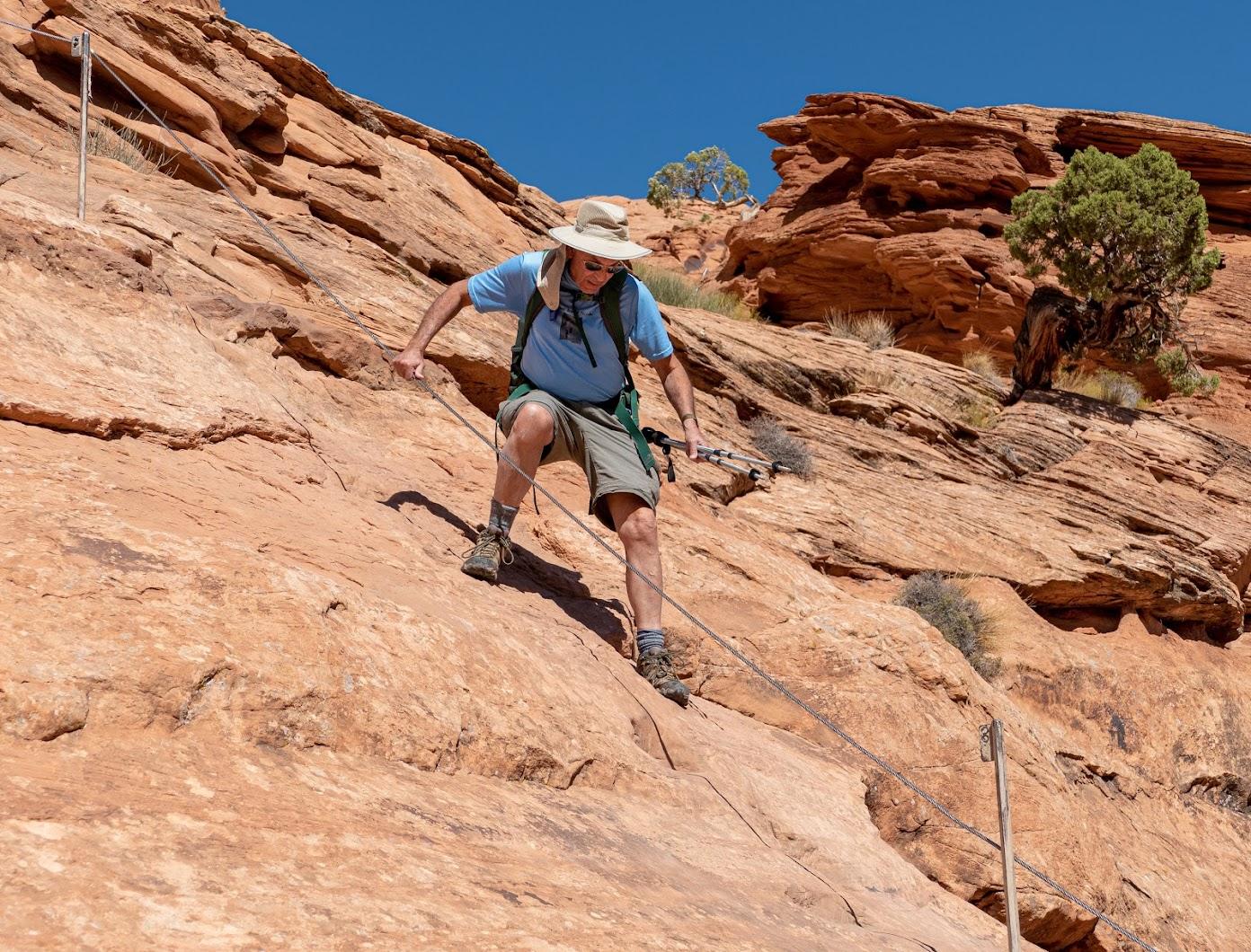 Man in hat and backpack carefully descends a steep red rock slope using ropes.