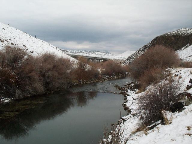 Snowy valley with a river, bare bushes, and a distant bridge under cloudy skies.