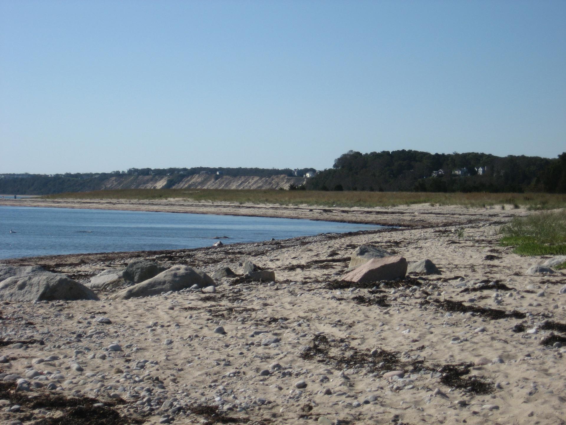 Sandy beach with rocks and distant dunes under a clear blue sky.