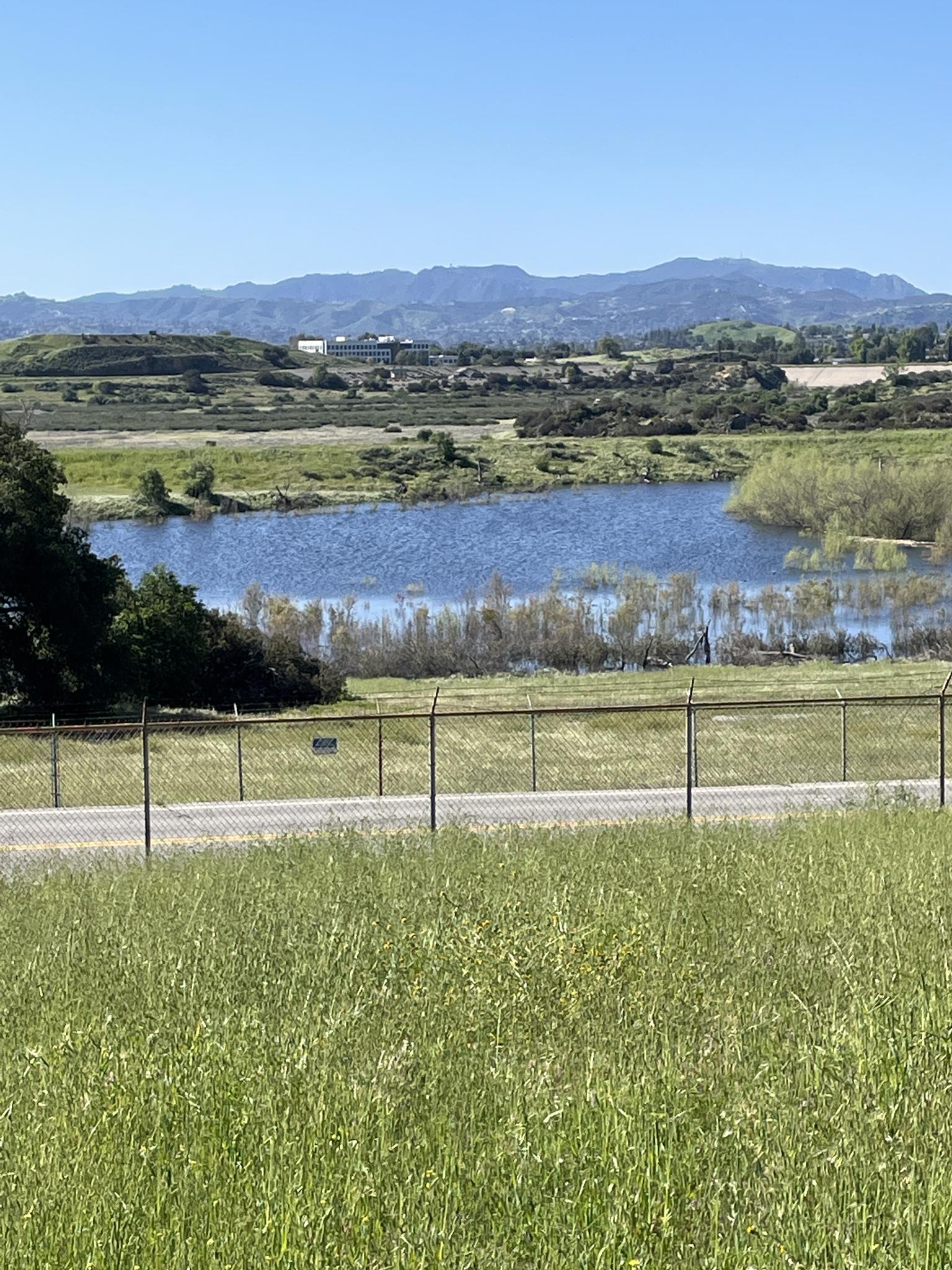 Grassy field and fence overlooking a lake with distant mountains under blue sky.