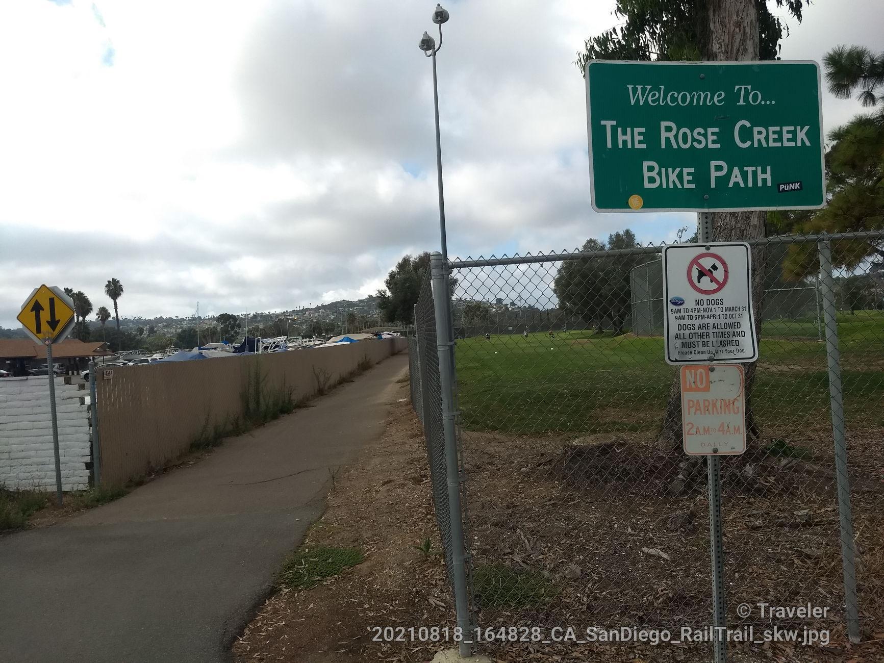 A green sign welcomes to a bike path next to a fence and paved trail.