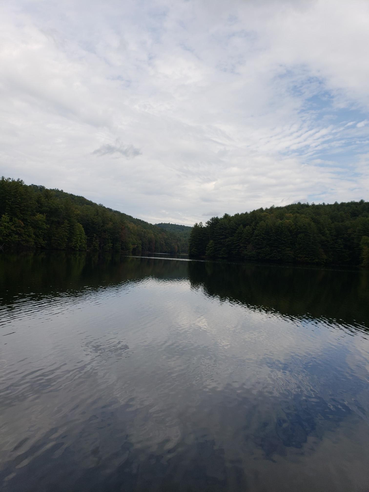Calm lake reflecting forested hills under a bright, cloudy sky.