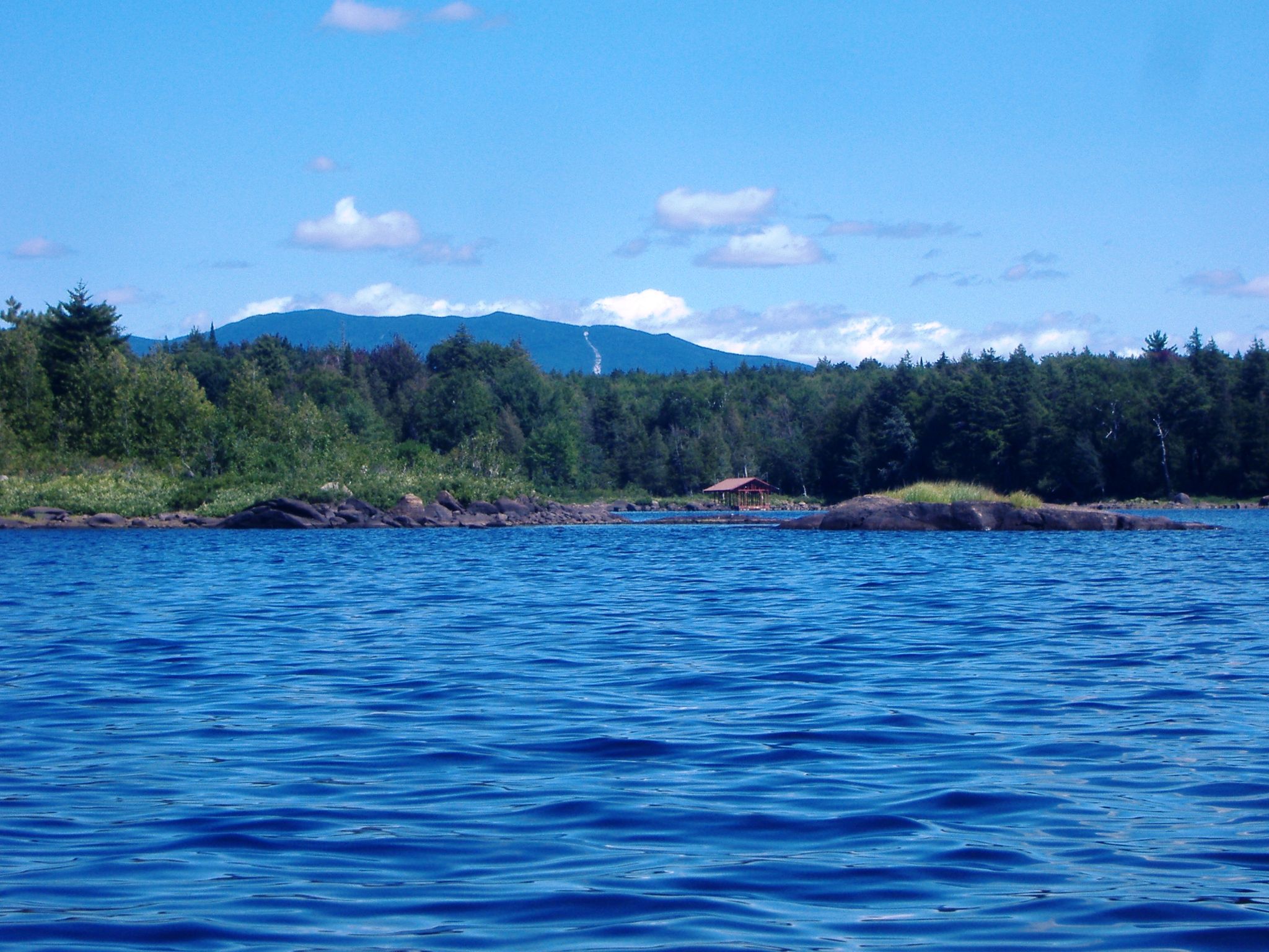 Vast blue lake with forested shore, distant mountains, and a small cabin. - Long Lake via Northville-Placid Trail