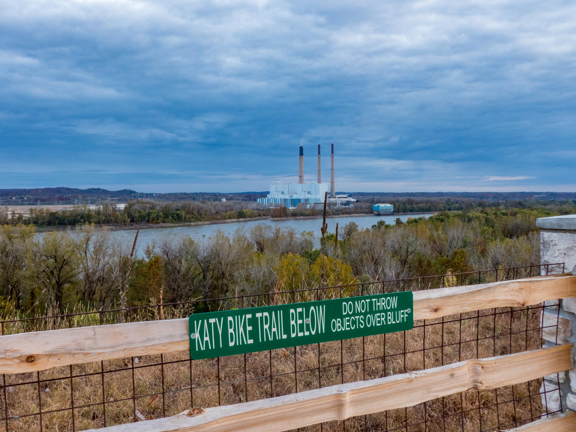 Overcast view of a river, industrial plant, and a fence with a trail sign.