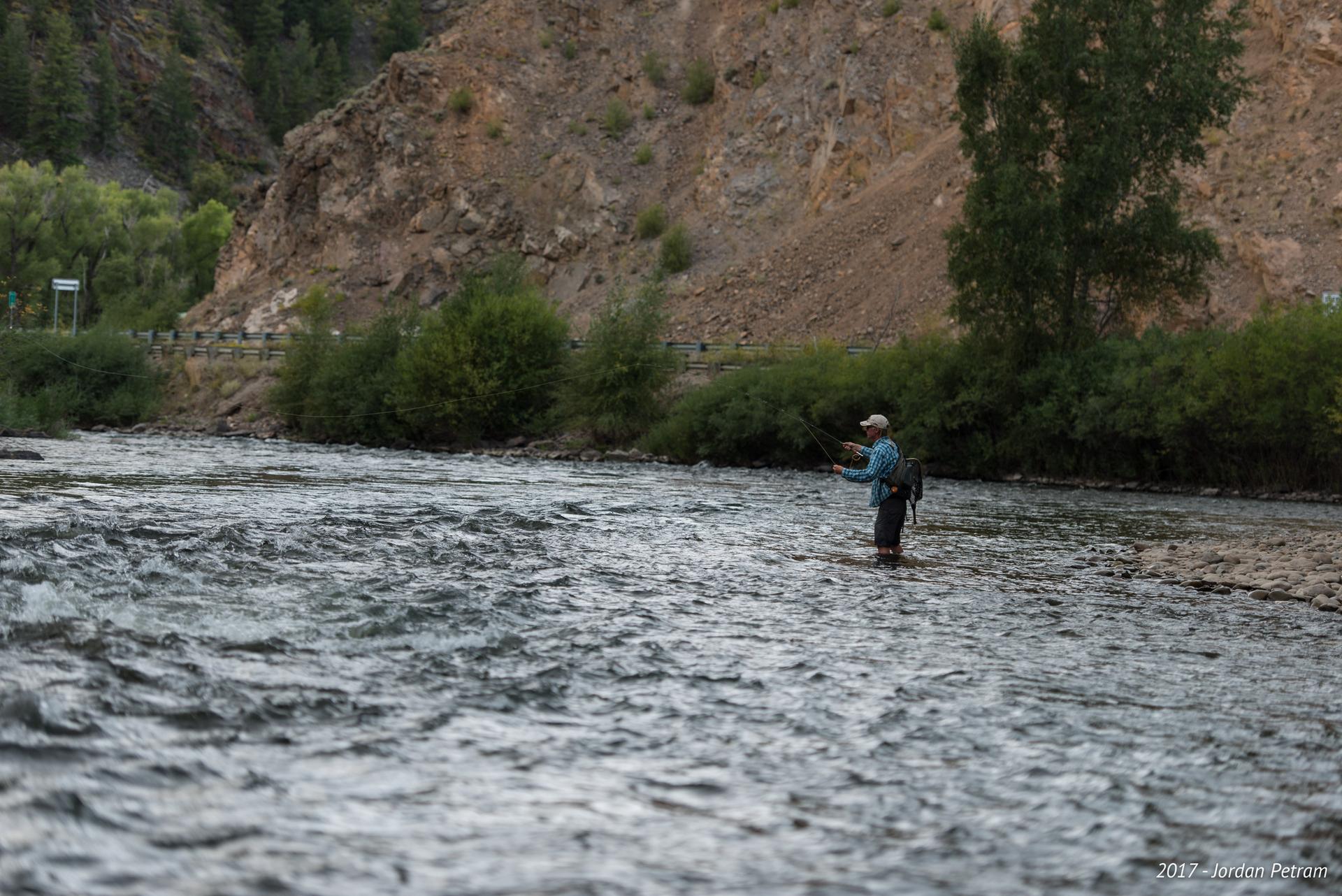 Fly fisherman wades in a river with a steep, rocky bank.