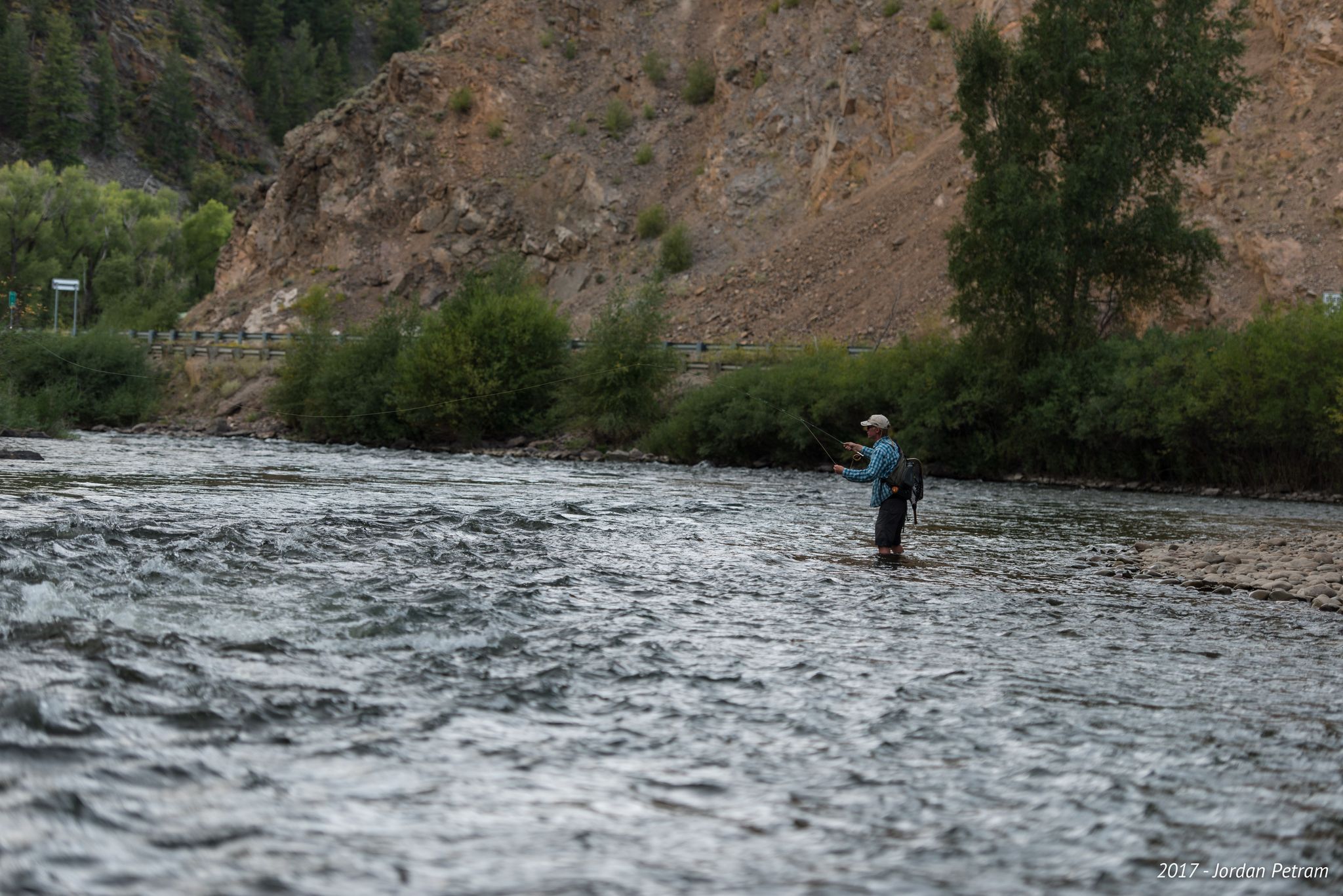 Fly fisherman wades in a river with a steep, rocky bank. - Almont to Jacks Cabin Cutoff Road gravel