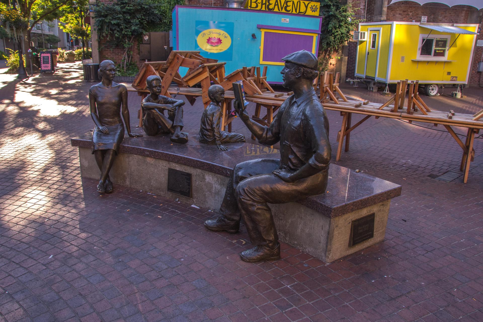 Bronze statues of people sitting on a bench in a sunny outdoor plaza.