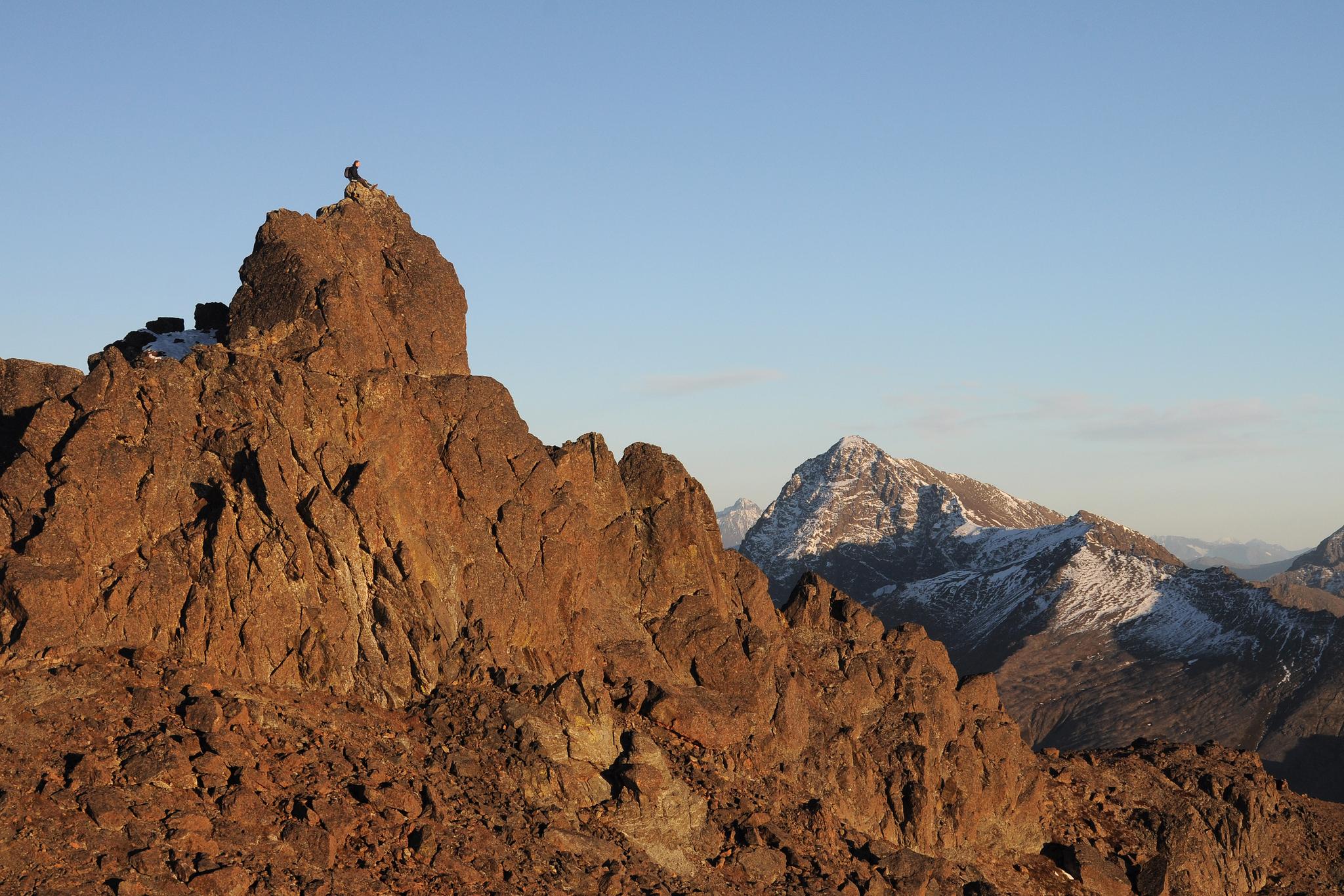 Person on a rocky peak overlooking snow-capped mountains under clear sky.