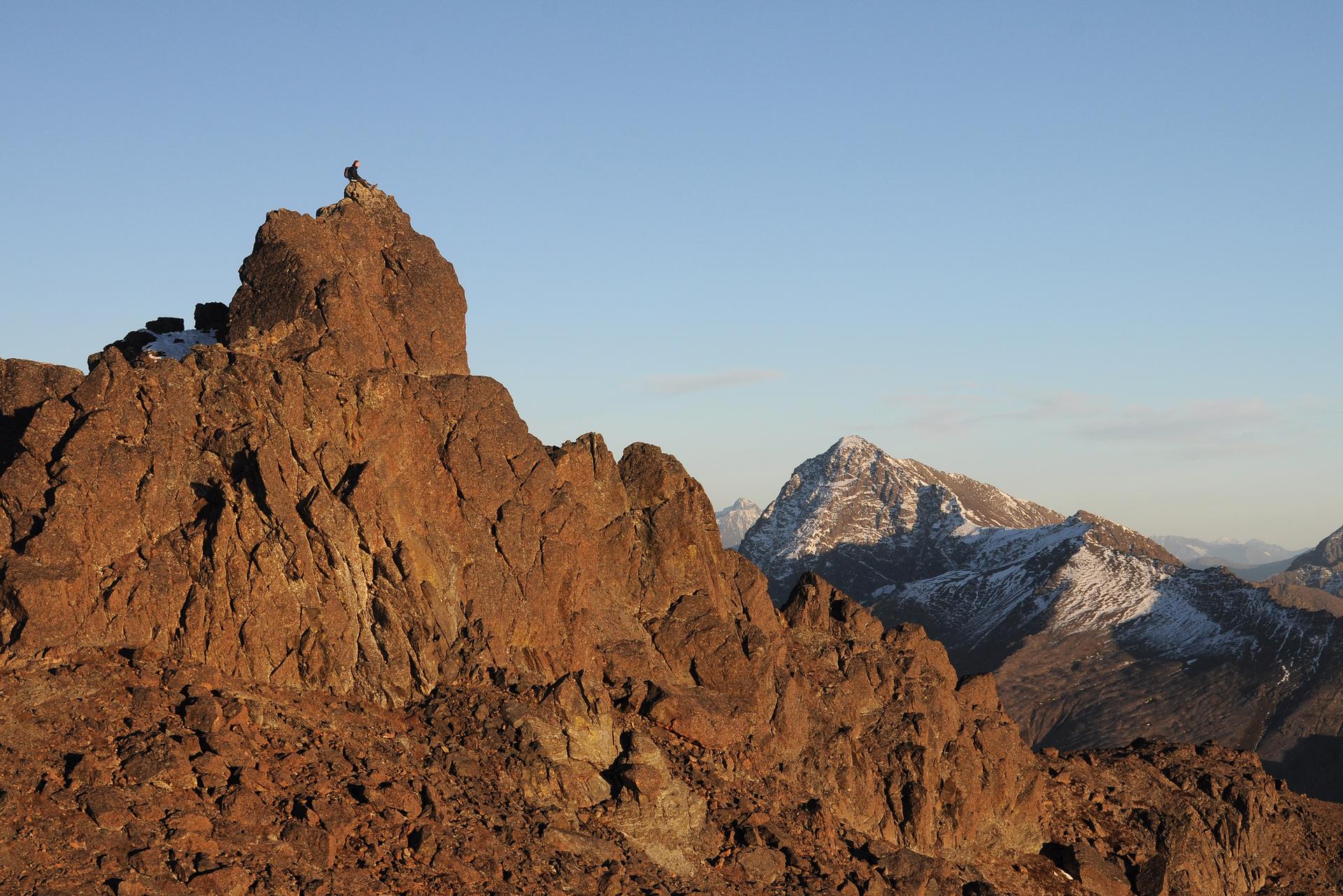Person on a rocky peak overlooking snow-capped mountains under clear sky.