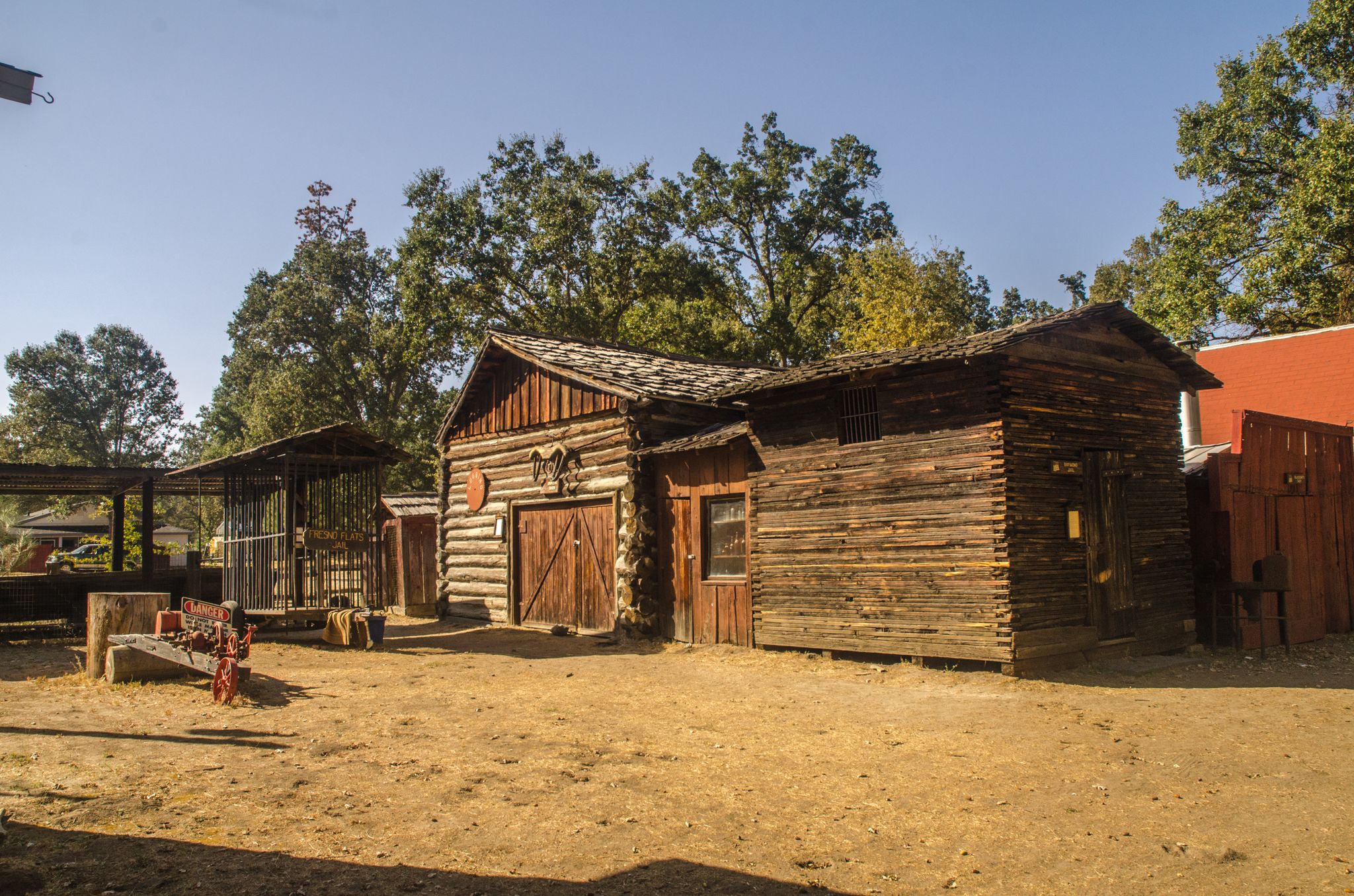 Rustic wooden buildings in a dirt yard, surrounded by trees on a sunny day. - Oakhurst, California to Wassama Round House State Historic Park