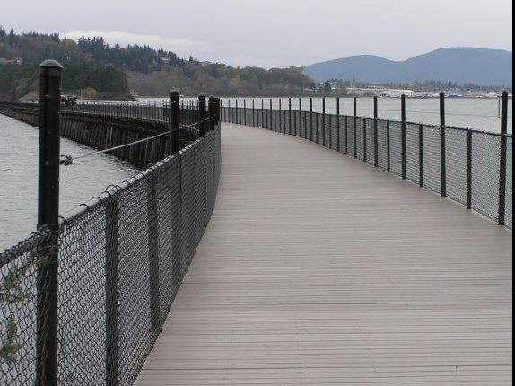 Long boardwalk with chain-link fences extending over water, overcast day.