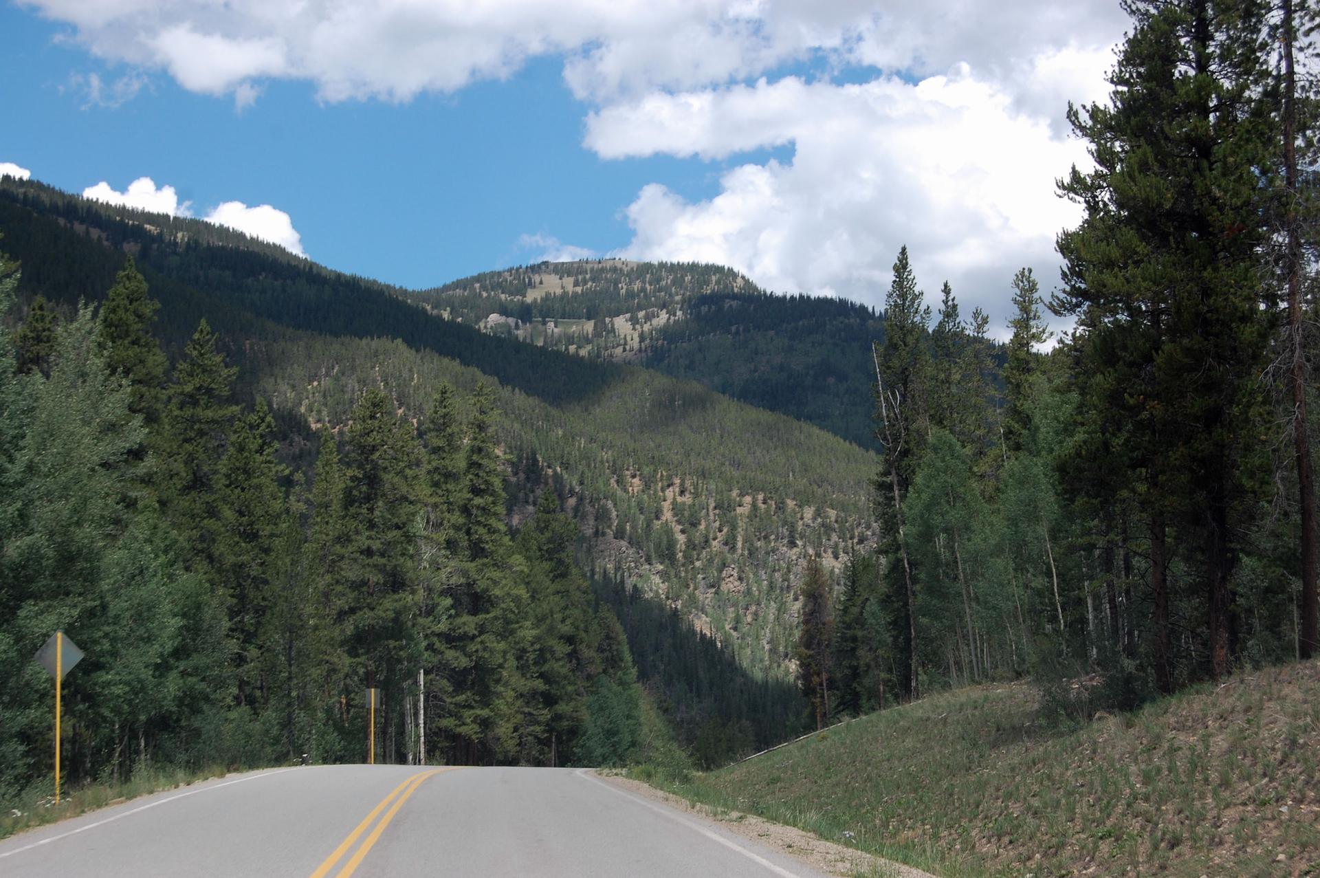 Road through dense green mountains under a bright, cloudy sky.