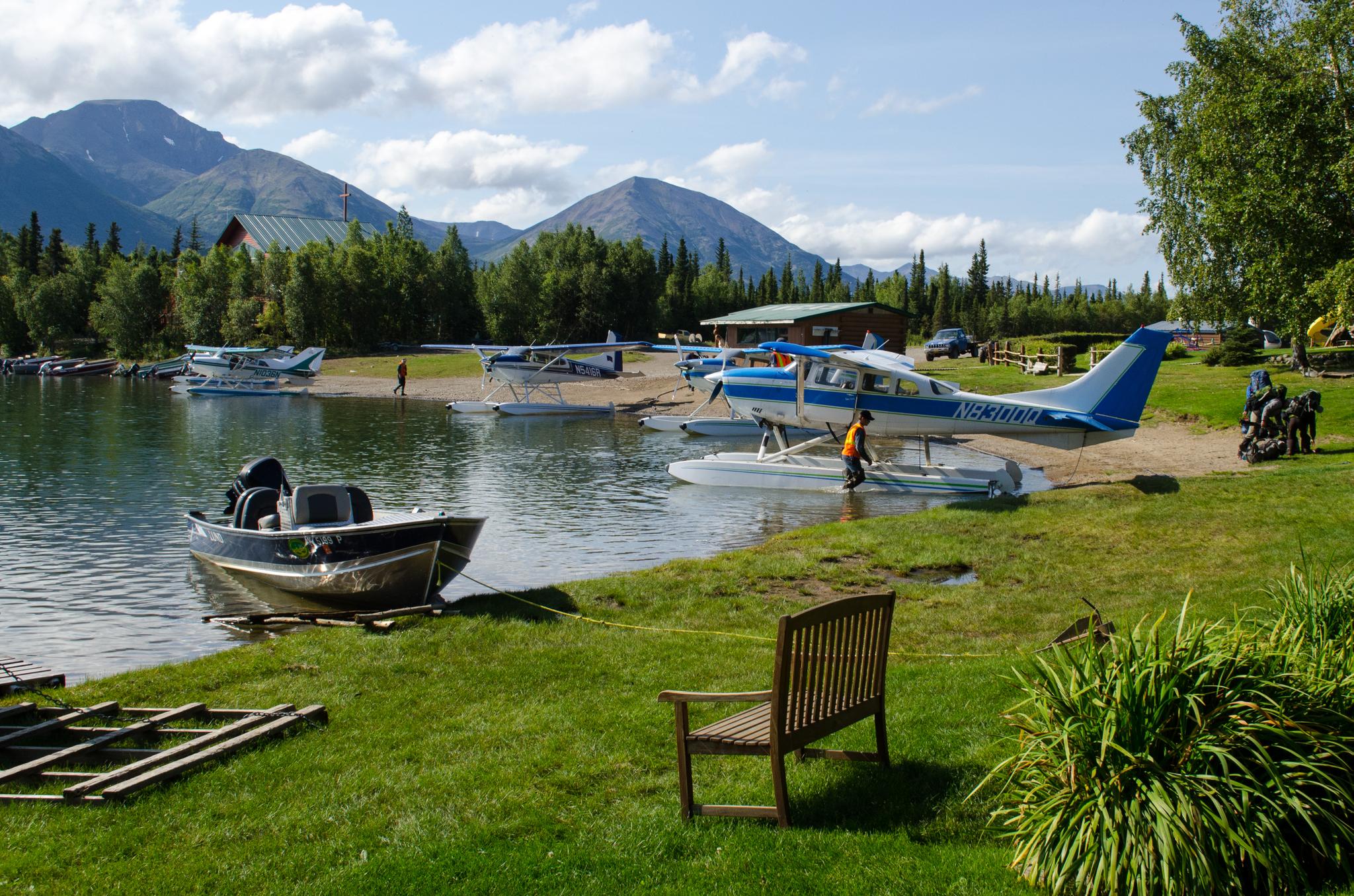 Seaplanes and a boat on a lake shore with mountains under sunny skies.