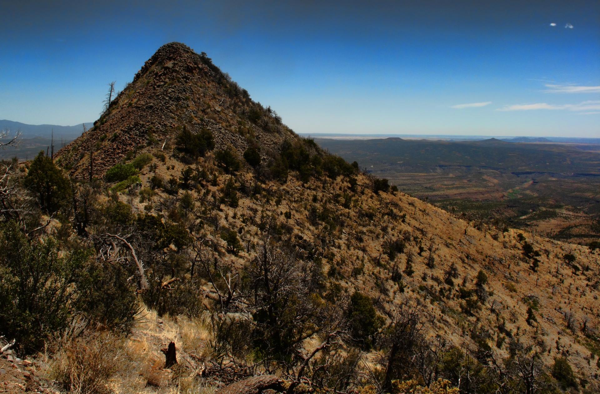 A rugged, conical mountain peak under a clear blue sky, overlooking arid plains.