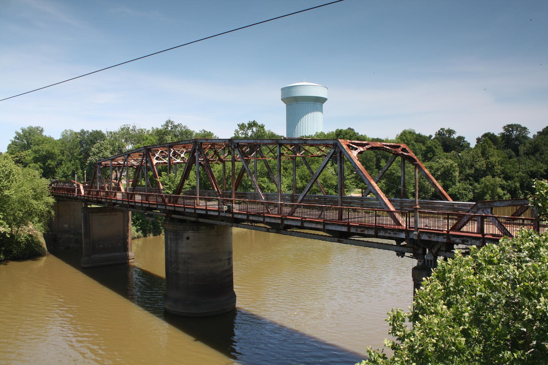 Sunny view of a rusty metal truss bridge over a brown river.