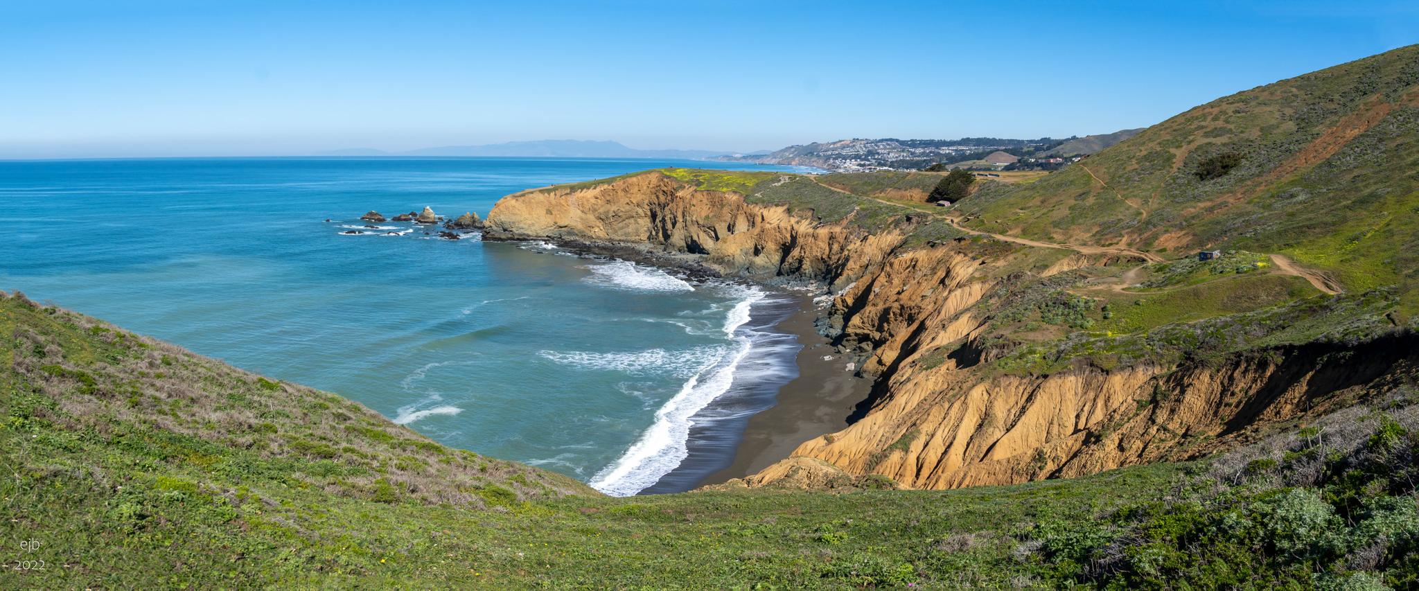 Sunny coastal view with rugged cliffs, blue ocean, and green hills.