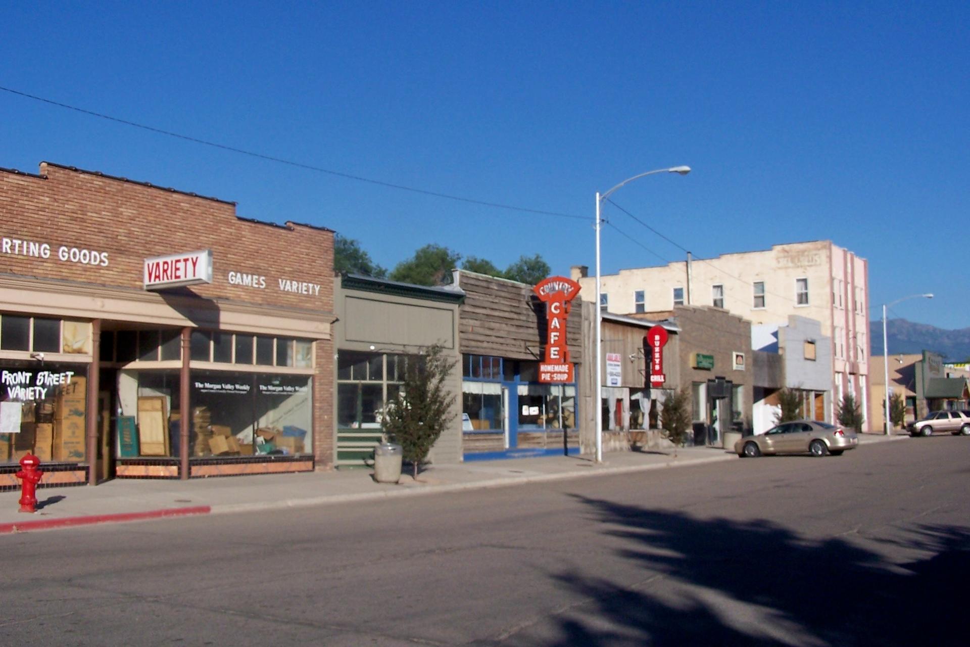 A sunny street with brick buildings, storefronts, and parked cars under a clear sky.