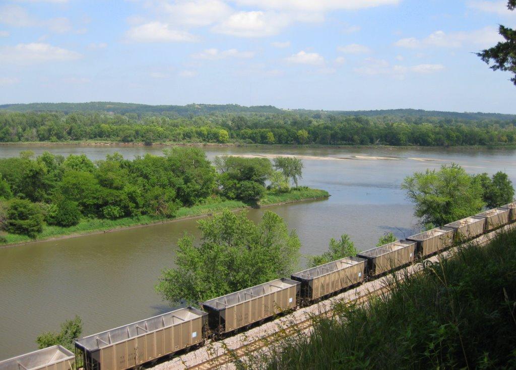 Long train cars on tracks beside a wide river with green trees.
